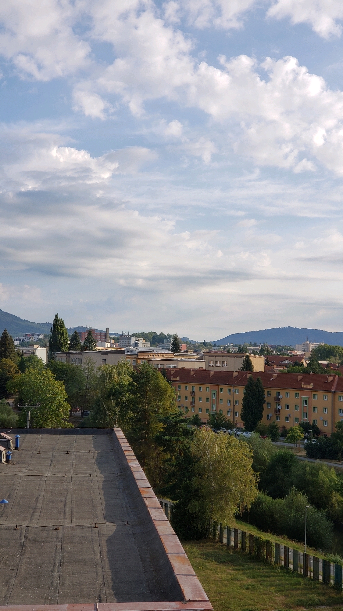 This is a high-angle, vertical, full shot of a cityscape in Prešov, Slovakia, under a partly cloudy sky. In the foreground, the flat, dark roof of a building with visible seams and some debris is prominent. A weathered brick parapet runs along the edge of the roof. Beyond the roof, a dense line of lush green trees forms a visual barrier, obscuring much of the ground level.
Further into the mid-ground, a cluster of apartment buildings, predominantly yellow and orange with red roofs, are visible. These buildings have multiple stories with numerous windows and balconies. Some of the apartment buildings are partially obscured by trees. Beyond the apartment buildings, other urban structures, including taller buildings with windows and darker facades, can be seen. In the background, rolling hills or mountains form the horizon. The sky above is a pale blue, interspersed with fluffy white and grey clouds, some of which are illuminated by sunlight, suggesting it is either morning or late afternoon. The overall impression is one of a residential or mixed-use urban area integrated with natural greenery, possibly on the outskirts of the city, given the presence of hills.