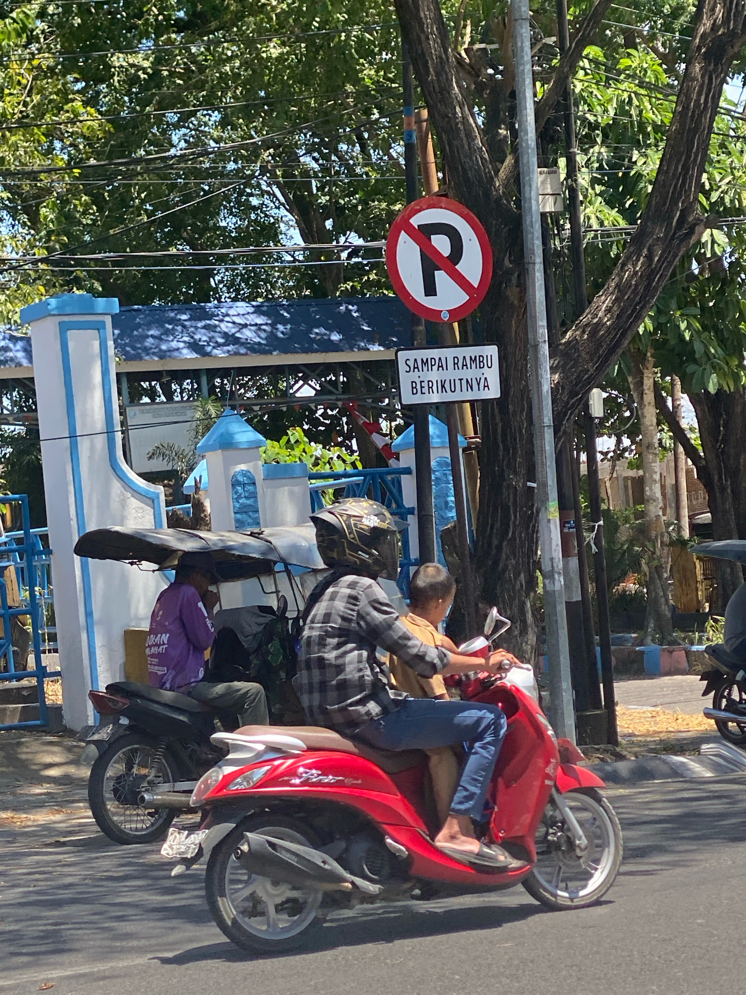 This is a medium shot, eye-level photograph taken in Kalumeme, Indonesia, under a clear blue sky. The scene depicts a street with several motorbikes and a tri-motorcycle. In the foreground, a red scooter is ridden by a person wearing a plaid shirt and blue jeans, with another person in a tan shirt behind them. To the left, a black motorbike is ridden by a person in a purple shirt, with a person wearing a helmet and black jacket behind them. A tri-motorcycle with a black canopy is also visible. Traffic signs are present, including a "No Parking" sign and another sign with Indonesian text. The environment includes trees with lush green foliage, overhead power lines, and a blue and white building in the background. The street is paved, with some dry leaves scattered on the surface. The overall impression is a typical street scene in Indonesia on a sunny day.