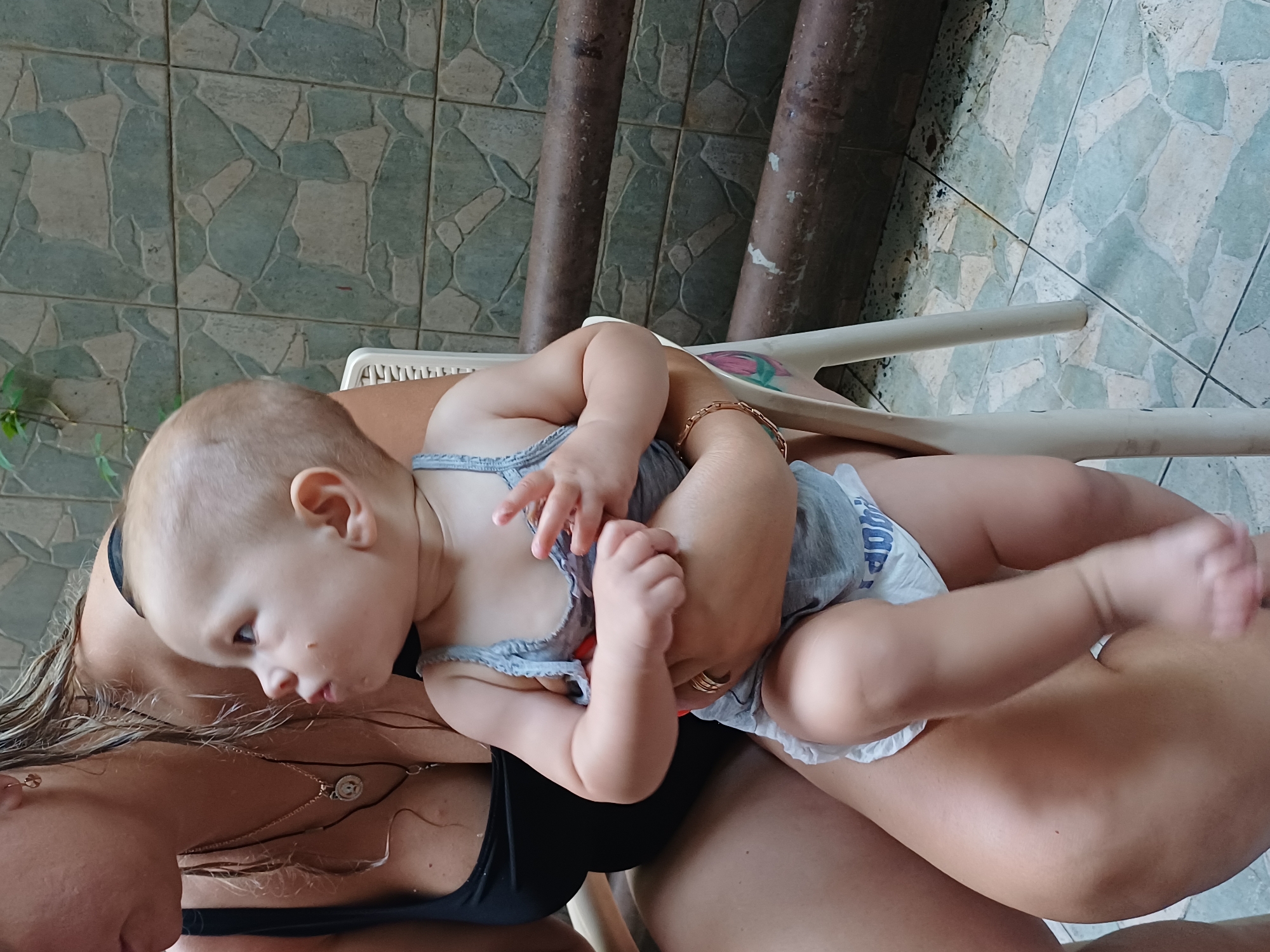 In this image, a woman is holding a baby in her arms. The baby is wearing a grey top and a diaper, and is looking to the side with a curious expression. The woman is wearing a black bikini top and a necklace. The scene appears to be outdoors, with a tiled floor and some greenery visible in the background. The lighting suggests it is daytime. The woman is holding the baby on her lap, and the baby's legs are extended. A bracelet is visible on the woman's wrist. The overall impression is a tender moment between a mother and her child. The image was taken in Sochi, Russia.