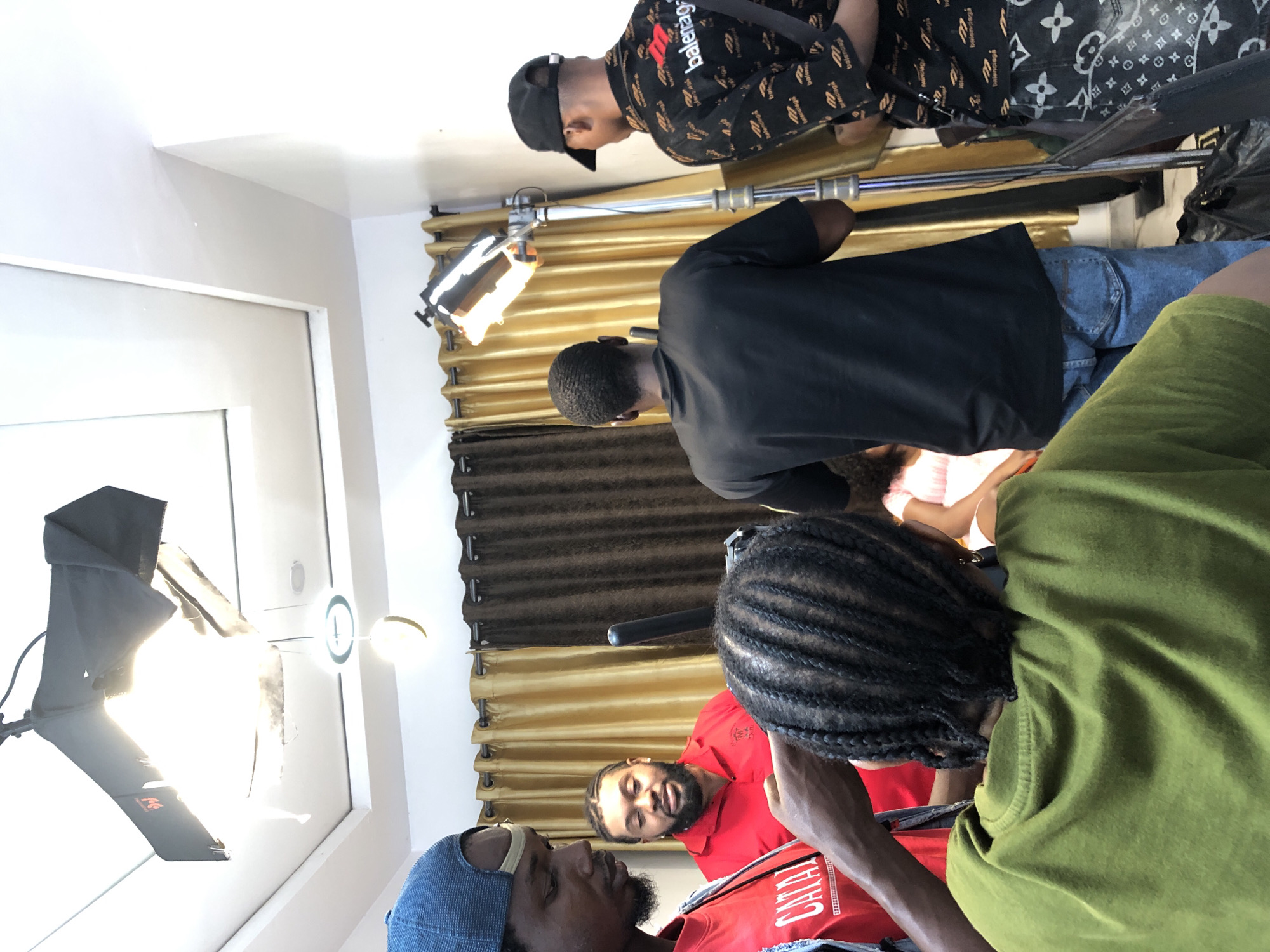 This image depicts an indoor scene, likely a film or recording set, featuring at least six individuals and professional lighting equipment. The setting is a room with white walls and ceiling, and prominent brown and gold curtains acting as a backdrop.
In the upper left, a man wearing a black cap and a patterned black shirt adorned with "Balenciaga" branding is visible, looking downwards. Below him, another man in a plain black long-sleeved shirt and blue jeans stands, facing away from the camera. To the right of this man, partially obscured, is a person in a green top with braided hair, also facing away.
Towards the center and lower part of the frame, three men are seated or leaning. One man in a red shirt with a beard looks up and to the left, displaying a slight smile. In the lower left foreground, another man wearing a blue cap and a red shirt with the text "CATALAYA" is visible. He also has a beard and looks towards the right. A partially visible person in a light-colored top is seated further back on the right.
The scene is illuminated by artificial lights, including a large light panel or softbox on a stand emitting a strong white glow on the left, and a smaller, warmer-toned light source on a stand in the background. A circular ceiling light fixture is also present. The presence of multiple people and professional lighting suggests a production or collaborative working environment, such as a video shoot, interview, or a similar creative project. One individual's jeans feature a distinctive Louis Vuitton monogram pattern.