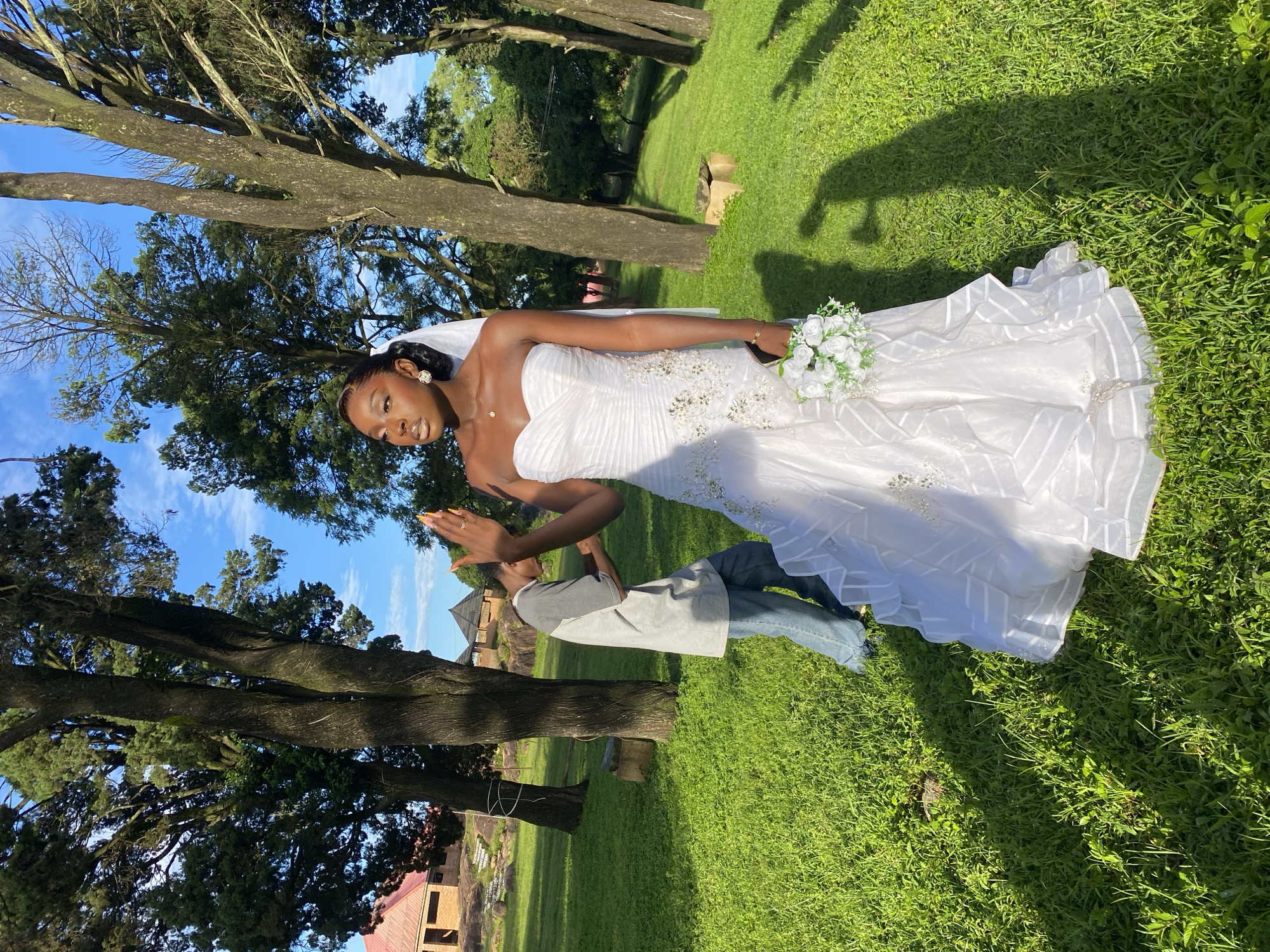 A young Black woman, dressed in a strapless white wedding gown with intricate beading and ruffled layers, stands in a grassy outdoor area. She holds a small bouquet of white flowers in her left hand and raises her right hand, with manicured nails, as if waving. She wears earrings and a necklace. Behind her and to the left, a person wearing a grey t-shirt and blue jeans bends over, appearing to adjust something. The setting is lush green grass with large trees and some buildings in the background, suggesting a rural or semi-rural location. The sky is blue with scattered clouds. The overall impression is a wedding photo shoot in a natural, sunlit environment. The location is described as Jos, Nigeria.