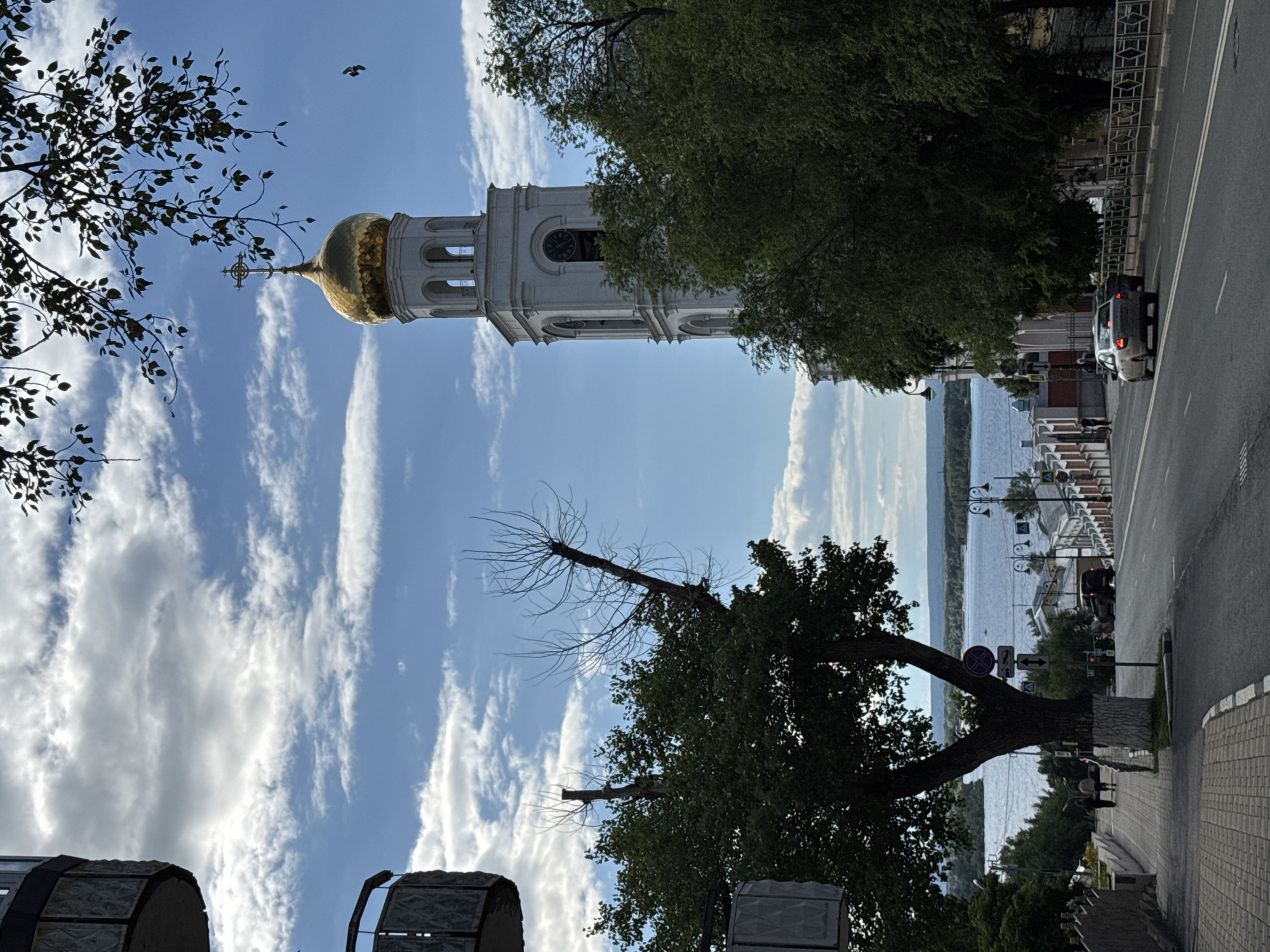 The image depicts an outdoor scene of a white bell tower with a golden dome topped with a cross, set against a bright blue sky with scattered white clouds. The bell tower has multiple arched openings and appears to be part of a religious structure. In the foreground, there are trees with green foliage and bare branches. The scene is oriented vertically.
To the right of the bell tower, a street runs alongside a body of water, likely a river or lake, with trees and buildings visible on the opposite bank. A silver car is parked on the street, and a few pedestrians can be seen on the sidewalk. The overall setting suggests an urban environment near a waterfront. The lighting indicates it is daytime, with clear skies and sunshine.
The image composition, with the bell tower dominating the upper portion and the street and water in the lower right, creates a sense of depth. The presence of trees and the architectural style of the bell tower hint at a historical or cultural site. The image appears to be taken from a slightly elevated position, looking up towards the bell tower and down towards the street and water. No visible text is present in the image.