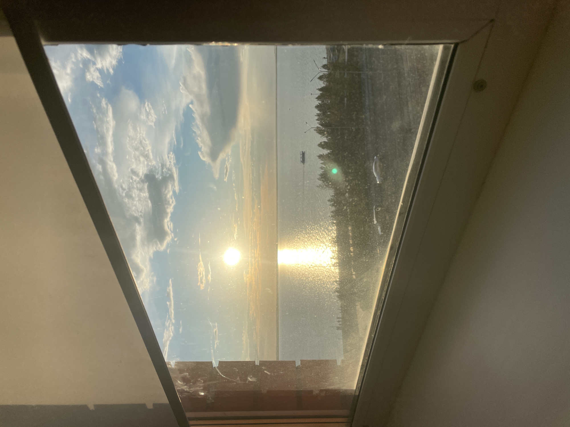 The image displays an exterior scene of a sunset over a large body of water, viewed through a dirty interior window. The window's light-colored frame is prominent, revealing smudges, streaks, and particulate matter on the glass, particularly on the left vertical and lower horizontal panes. The sun, a distinct bright circular disc, is positioned low on the horizon, slightly left of the image's vertical center, casting a brilliant golden columnar reflection across the calm water surface. The sky is clear blue with scattered white cumulus clouds, more concentrated in the upper right. A dark, narrow strip of land forms a distant shoreline, backed by a sparse line of dark green trees. Below the trees, a paved road runs parallel to the water's edge, occupied by two light-colored passenger vehicles. Multiple dark-colored streetlamp poles are interspersed among the trees. Several small human figures are discernible on the shoreline, with one individual clearly standing on a raised section on the right. A single dark-hulled boat is visible on the water to the right of the sun's reflection. The scene is located in Kokshetau, Kazakhstan, with the water body identifiable as Lake Kopa.