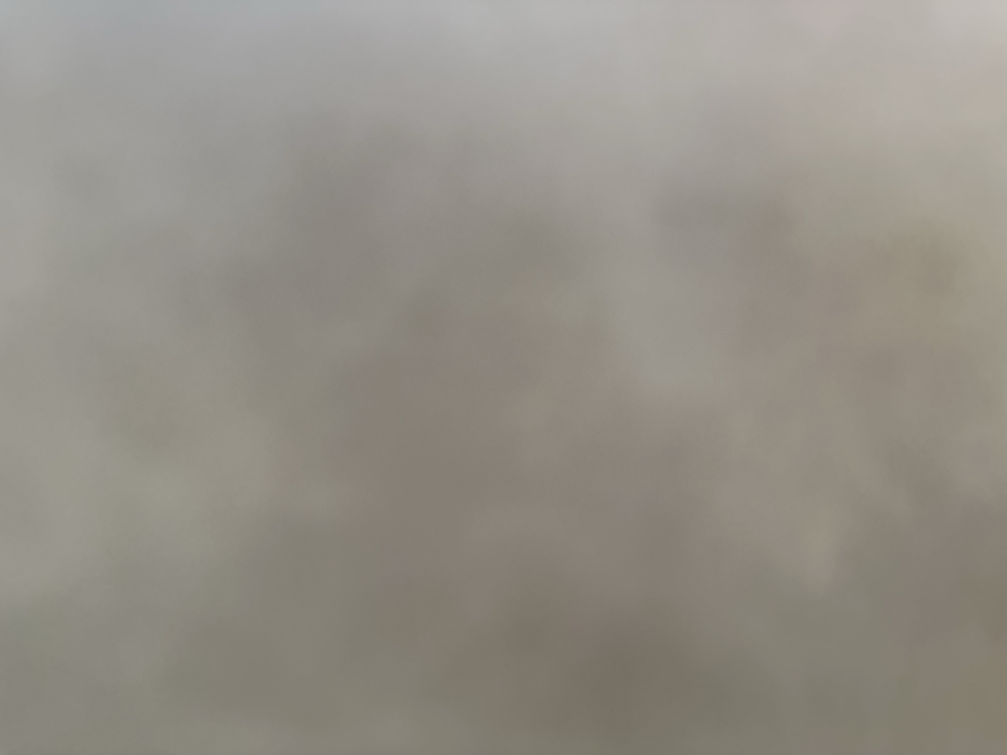 This is a low-resolution, blurry image with a predominantly grey and beige color palette. The texture appears to be uniform, suggesting a flat surface like a wall or fabric. There are no discernible subjects, objects, or scenes. The image lacks any clear details that would indicate a specific time of day, weather conditions, or location. No text is visible. The overall impression is that of an abstract or out-of-focus photograph.
