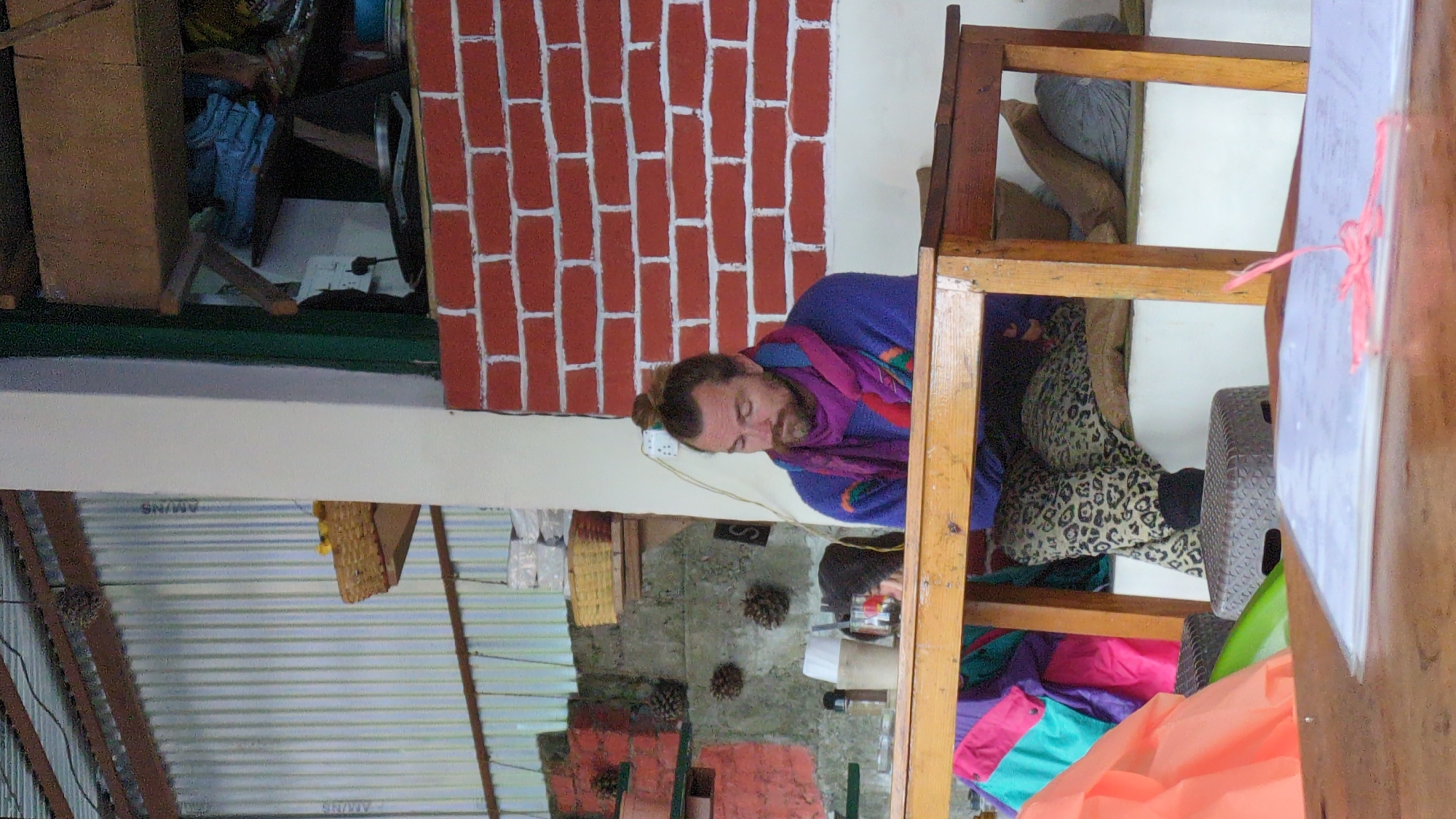 The image shows an interior scene. A man with a beard and hair pulled into a bun is wearing a blue shirt and a purple scarf, standing next to a wall with a red brick pattern. A wooden structure, possibly shelving or a table frame, is visible. Various objects and textiles are scattered around the structure, including a leopard-print garment, colorful fabrics, and a plastic stool. The wall behind him is light-colored. Below this area, the setting includes corrugated metal sheets, suggesting a simple, perhaps makeshift, construction. Pine cones are also visible on a lower wall section. The image does not contain enough details to verify if it is located in Haini, India.