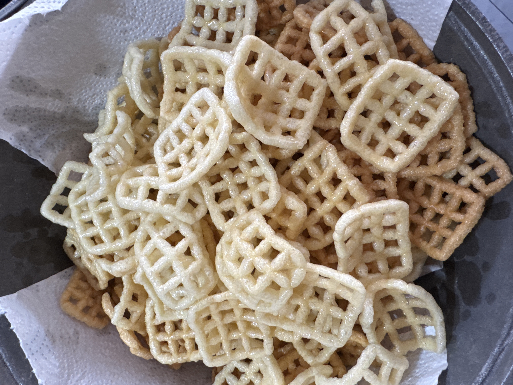 A close-up, overhead shot captures a pile of waffle-shaped chips resting on a paper towel inside a dark-colored dish. The chips are a light golden brown, with some appearing lighter and more translucent than others. The waffle pattern is clearly visible, with a lattice of interconnected squares. The chips are piled high, with many overlapping each other. Some of the chips appear to be slightly greasy, reflecting the light. The background is a soft white paper towel, with the dark dish visible at the bottom and edges of the image. The lighting is bright and diffused, creating a slightly hazy effect.