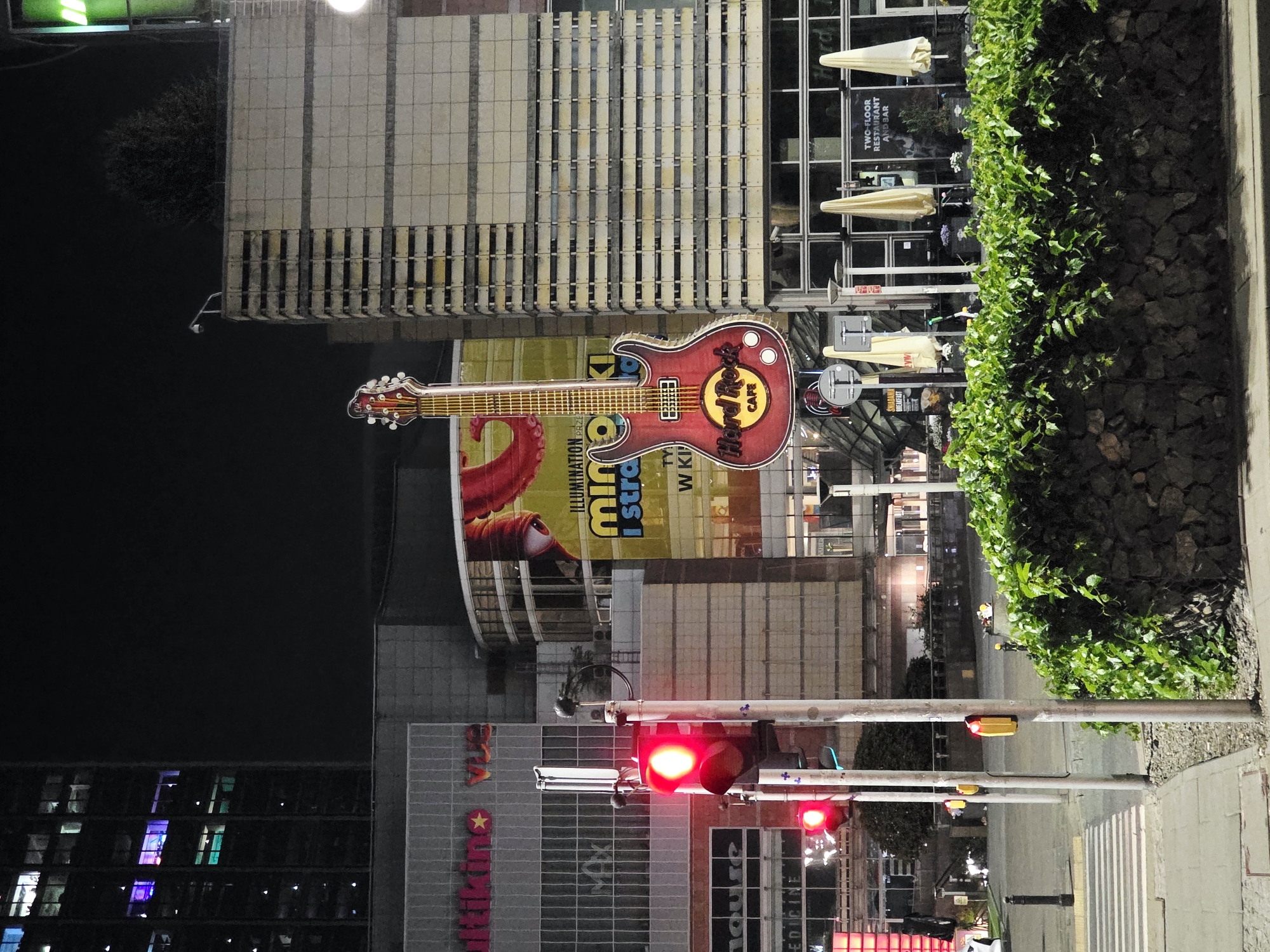 The image depicts a nighttime street scene in Warsaw, Poland, likely in a commercial district. A prominent, oversized red electric guitar sculpture with "Hard Rock Cafe" logo is displayed prominently. Buildings flank the street, with one on the left appearing to be a multi-story office building with illuminated windows showing various colors. Another building to the right is a lighter-colored facade with horizontal bands. In the foreground, a traffic light is illuminated red, indicating a stop. A crosswalk is visible on the left side of the image. A densely planted hedge with dark stone retaining wall runs along the right side of the street. Several outdoor umbrellas are visible on the right, suggesting an outdoor seating area. The overall atmosphere is urban and commercial, with the iconic Hard Rock Cafe guitar serving as a central landmark.