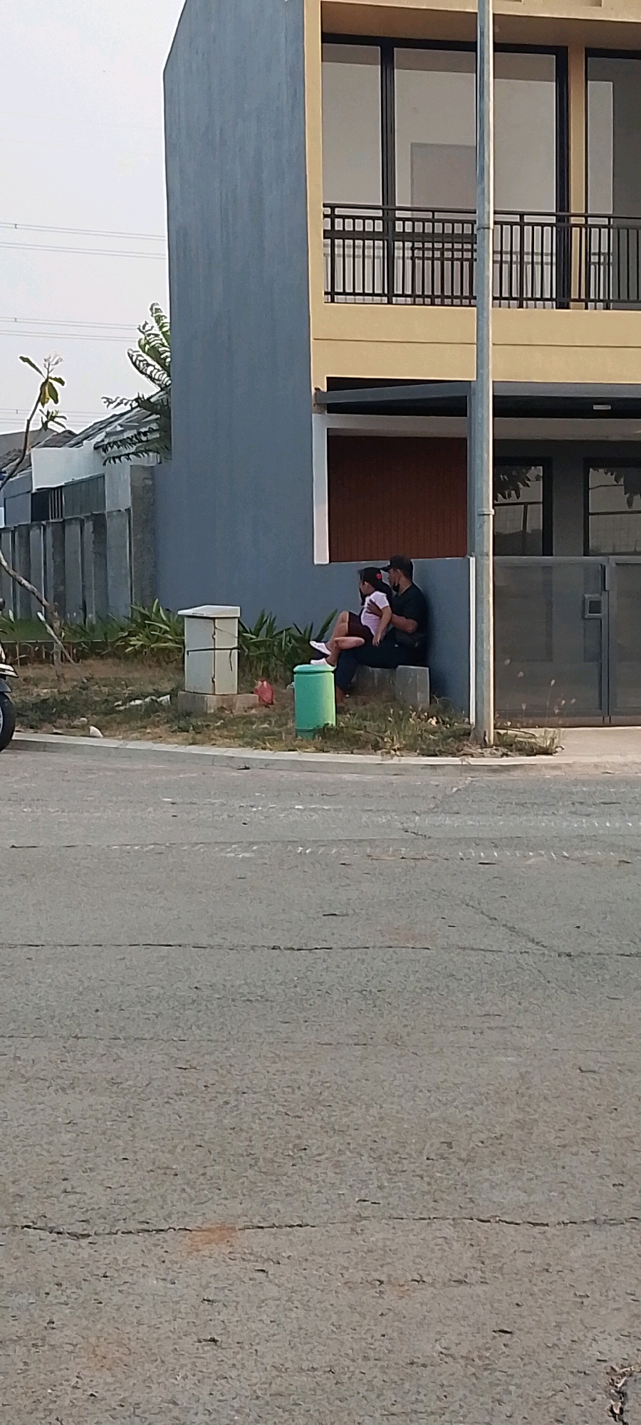 In Satriamekar, Indonesia, a man and a child are seated on a low concrete ledge in front of a modern house. The house has a gray plastered wall and a yellow upper story with large windows and a black metal balcony railing. The man, wearing a black t-shirt and a dark cap, holds the child on his lap. The child, dressed in a white top and maroon shorts, is facing the man. A green cylindrical trash bin is situated next to them, and a small red object, possibly a toy or a piece of trash, lies on the ground nearby. Behind the individuals is a section of the house with a dark brown textured wall, and to the left, a gray concrete fence and some vegetation are visible. The ground in front of the ledge is covered in dry grass and dirt, and a paved road is in the foreground. The sky is overcast.