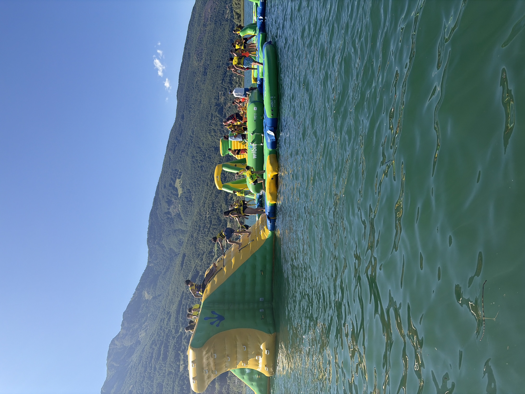 The image depicts an inflatable water park, branded with "Wibit," situated on a body of water, likely Lake Plav, in Montenegro. The park features various inflatable structures, including slides, climbing walls, and platforms in green, yellow, and blue colors. Several people, including adults and children, are actively engaged in climbing, sliding, and jumping on these inflatables. The water is calm and appears to be a deep green color. In the background, a large, tree-covered mountain range rises against a clear blue sky, typical of the mountainous landscape surrounding Plav. The scene conveys a sense of recreation and outdoor summer activity.