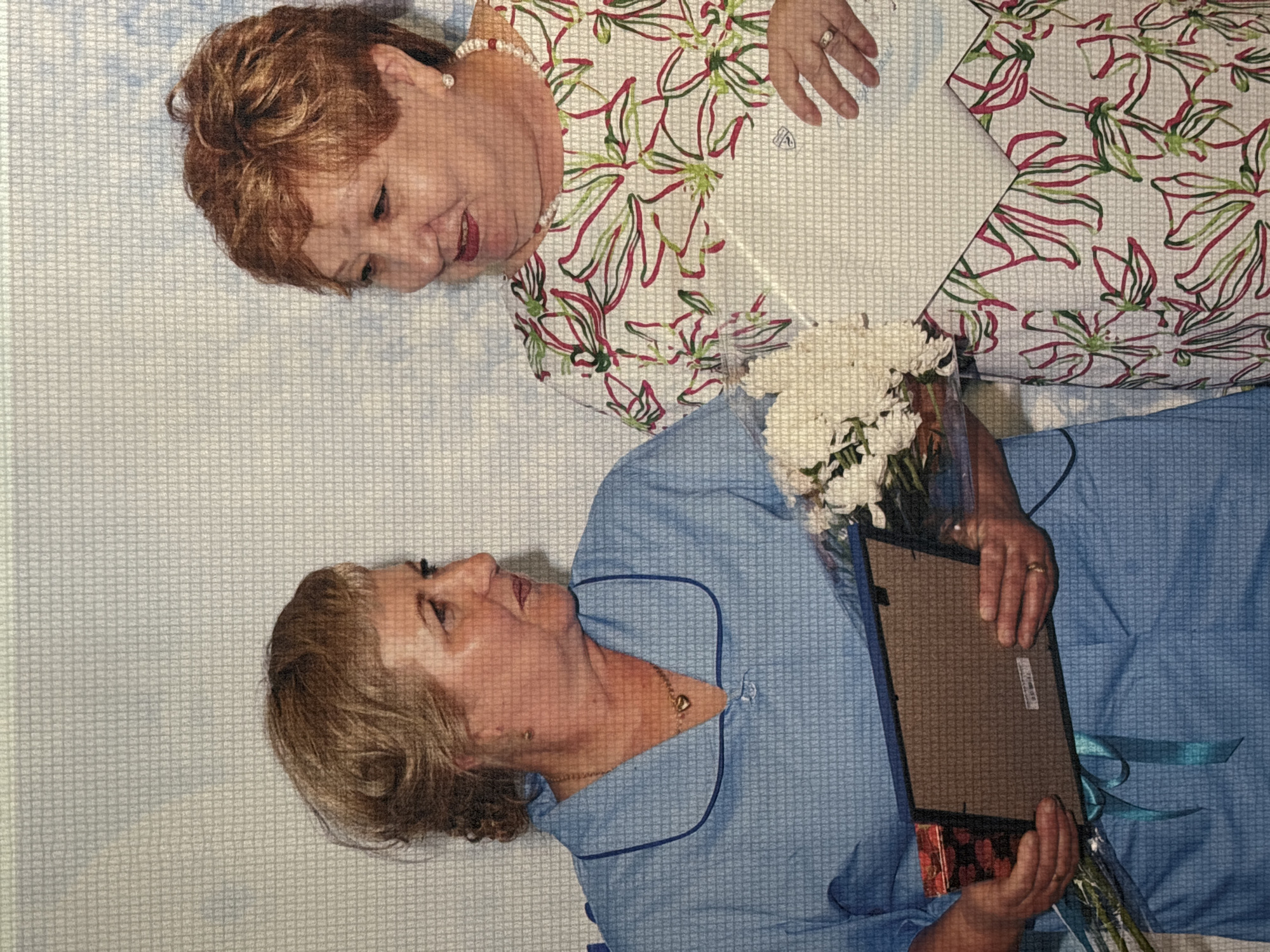 Two women are posing for a photograph in front of a textured, light-colored wall. The woman on the left, with short, light brown hair, wears a light blue uniform with a dark blue collar and a gold heart-shaped pendant necklace. She is holding a framed item and a bouquet of white flowers. The woman on the right, with short, reddish-brown hair, wears a white floral-print blouse with red and green outlines, and a pearl necklace. She is holding a document or certificate. Both women appear to be smiling and engaged in the photo opportunity. The image does not provide any specific clues to pinpoint a location within the city of Chaikovsky, Russia.
