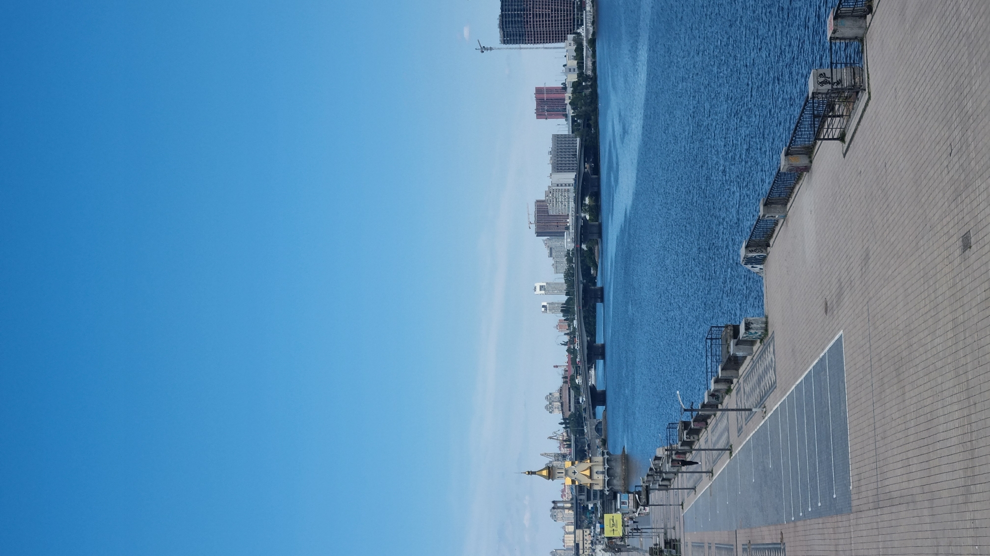The image depicts a wide, blue river with a paved embankment on the left. The embankment features parking spaces marked with white lines, lampposts, and decorative paving. A railing separates the embankment from the river. Across the river, a cityscape is visible, characterized by several bridges, modern buildings of various heights and architectural styles, and construction cranes. A prominent golden-domed church structure stands on a small island in the river on the left side of the image, near the embankment. The sky is a clear, light blue. The scene appears to be a waterfront view of Kyiv, Ukraine, likely overlooking the Dnipro River.