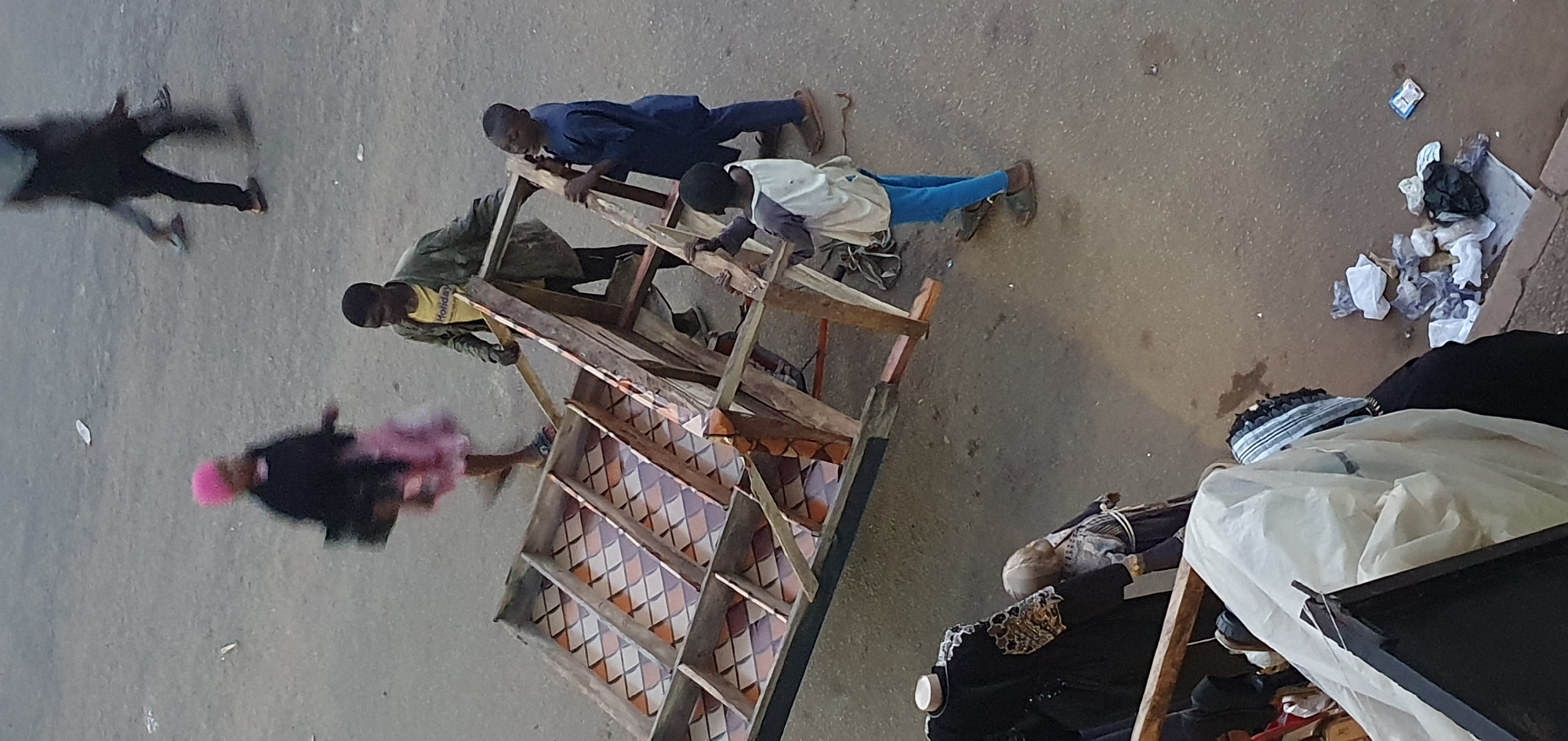 In an outdoor, urban setting on a dusty, paved surface, several individuals are engaged in moving a large, wooden structure. The structure appears to be a table or display unit with multiple shelves and a patterned inlay of orange and beige rhombuses. Three young men are actively involved in carrying the structure. One man in a blue outfit is on the right, gripping the side. Another in a green jacket and yellow shirt is in the middle, also holding the frame. A third, younger individual in a light-colored shirt and blue pants is on the left, supporting a section of the structure. In the background, a person with a pink headdress and a pink and black outfit walks away, blurred by motion. Another blurry figure is visible further in the distance. To the left of the frame, mannequins dressed in dark clothing with ornate trim are displayed. The ground is littered with various pieces of trash, including crumpled white and black plastic bags. The lighting suggests it is daytime.