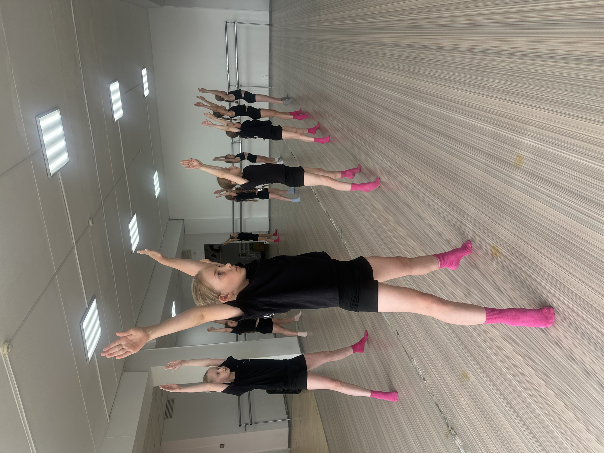 In an indoor dance studio located in Votkinsk, Russia, a group of young girls is engaged in a stretching exercise. The girls are aligned in a row, stretching with their arms extended horizontally. They are wearing black shorts, black shirts, and pink socks. The studio has a mirror on one wall, which reflects the scene. The floor is composed of light-colored stripes. The ceiling features rectangular lighting fixtures.