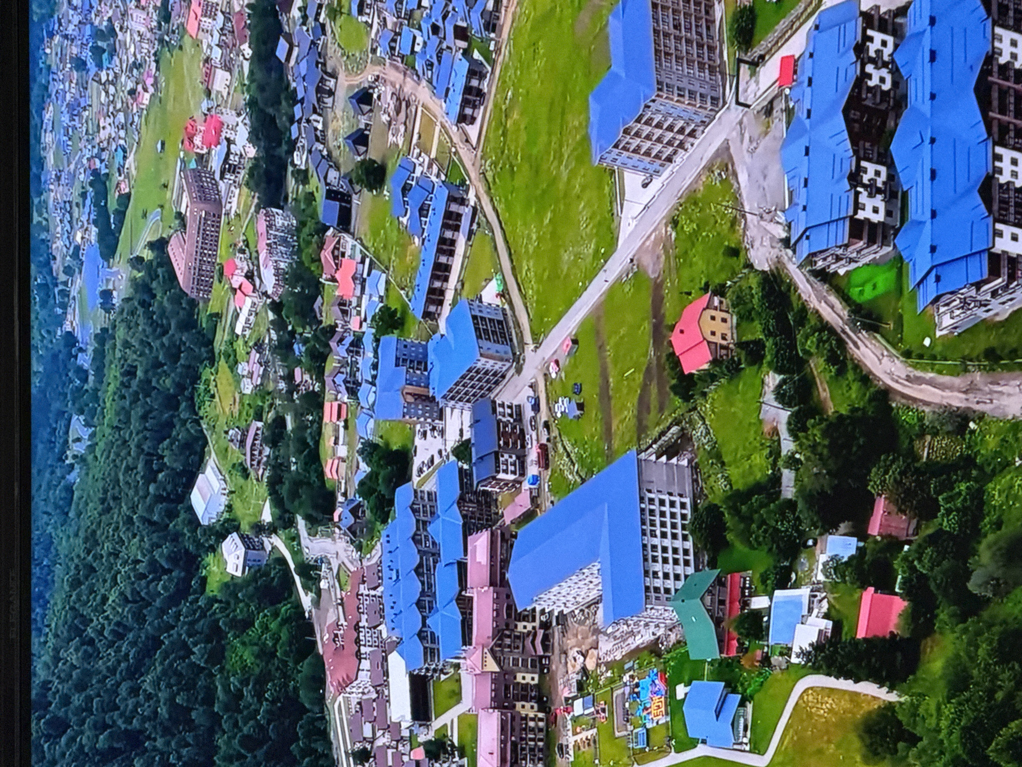 An aerial, eye-level, medium shot captures a mountain resort village. The scene is dominated by buildings with blue roofs and surrounding greenery. In the foreground, two large apartment buildings with intricate balcony designs and blue roofs are visible. Adjacent to these, a construction site is apparent with some building materials and excavation. To the left, smaller buildings with red and blue roofs dot the landscape, some appearing to be residential homes. A playground area with colorful inflatable structures is situated on the left side. Further in the background, more apartment complexes with blue roofs extend into the distance, interspersed with smaller houses and patches of forest. The environment appears to be a mountainous region, with a dense forest covering the slopes in the upper left portion of the image. A winding road is visible, connecting various parts of the village. The overall impression is that of a developing resort community nestled within a natural landscape.