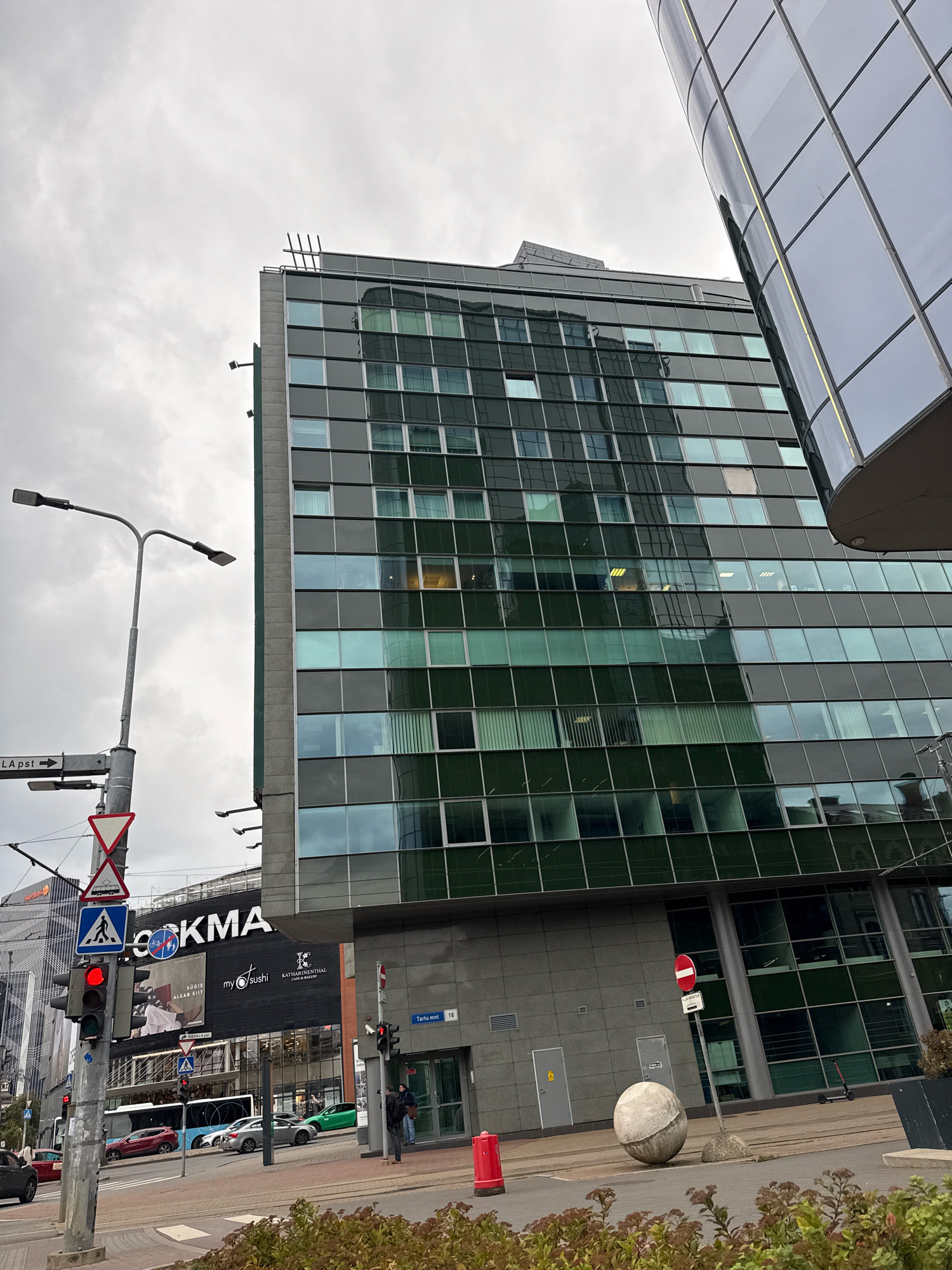This is a street-level, eye-level, medium shot of a modern cityscape in Tallinn, Estonia, under an overcast sky. The primary subject is a tall, multi-story building with a facade composed of dark green and reflective glass windows, reflecting the cloudy sky. Adjacent to it, another building with a curved, mirrored glass exterior is partially visible in the upper right corner, leaning into the frame.
In the foreground, a street scene unfolds. A traffic light with a red signal is prominent on the left, positioned on a metal pole with multiple traffic signs. One sign points to "LA PST" with an arrow. Below the red traffic light, a blue pedestrian crossing sign is displayed. A street lamp with two lights extends from the same pole.
Further down the street, a bus is visible, along with several cars. To the right of the street, a modern building with a dark facade displays large white lettering that reads "COCKMAN" and "my sushi." A cafe called "KATHARINENTAL CAFE & BAKERY" is also advertised.
On the sidewalk, a person in dark clothing is walking towards the right, near a building entrance. A large, spherical concrete sculpture rests on the pavement to the right of the person. Red bollards are present along the street. A scooter is parked near a planter in the background. The ground is paved, and there are some low-lying plants with brown and green foliage in the immediate foreground. The overall impression is a bustling urban environment on a cloudy day.