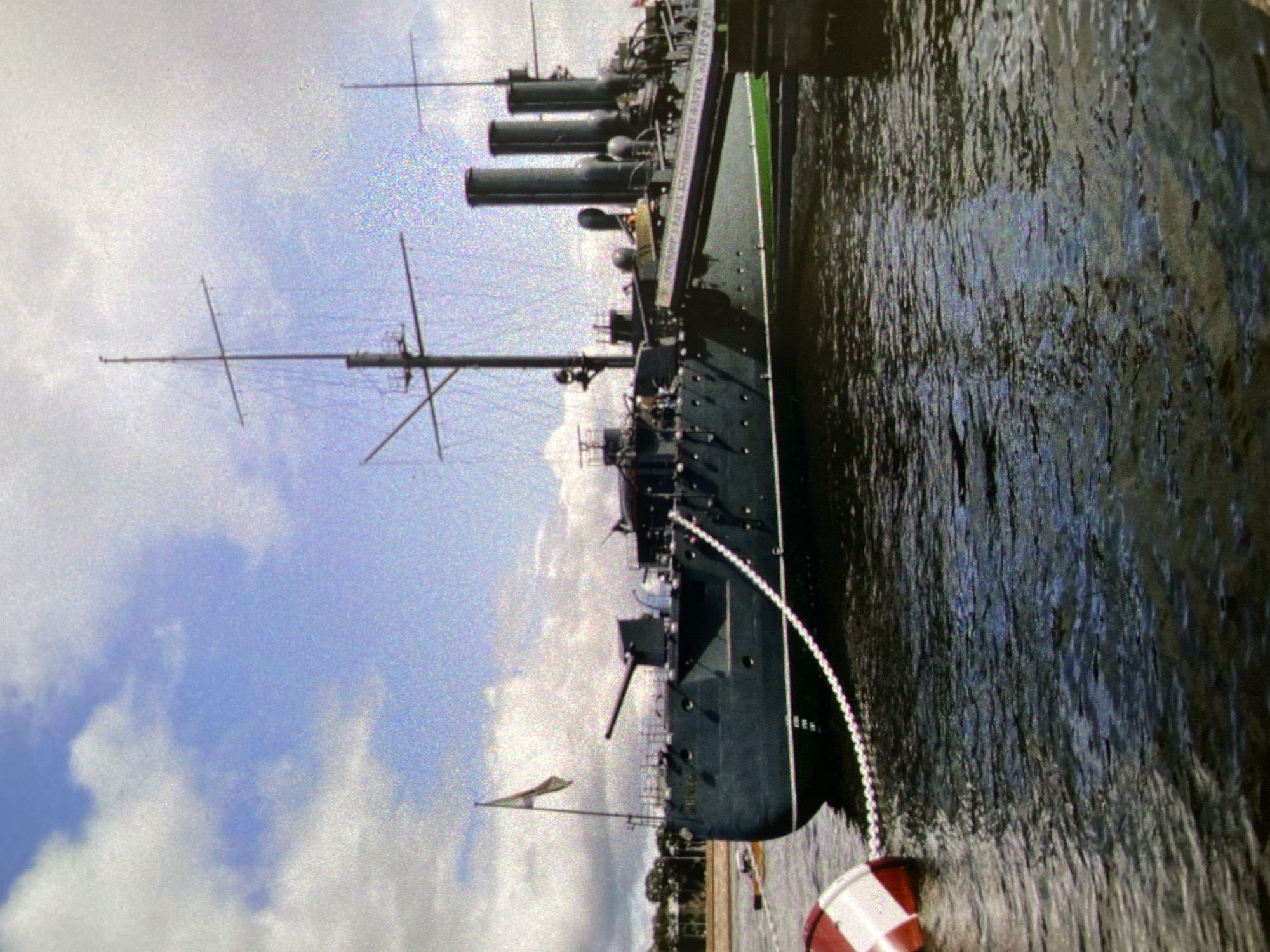 The image depicts the Russian warship Aurora, a historic cruiser docked in the Neva River in Saint Petersburg, Russia. The ship is dark gray, with several funnels and masts. The sky is partly cloudy. The water is dark and rippled. A red and white buoy is visible in the foreground. The ship is a museum ship.
