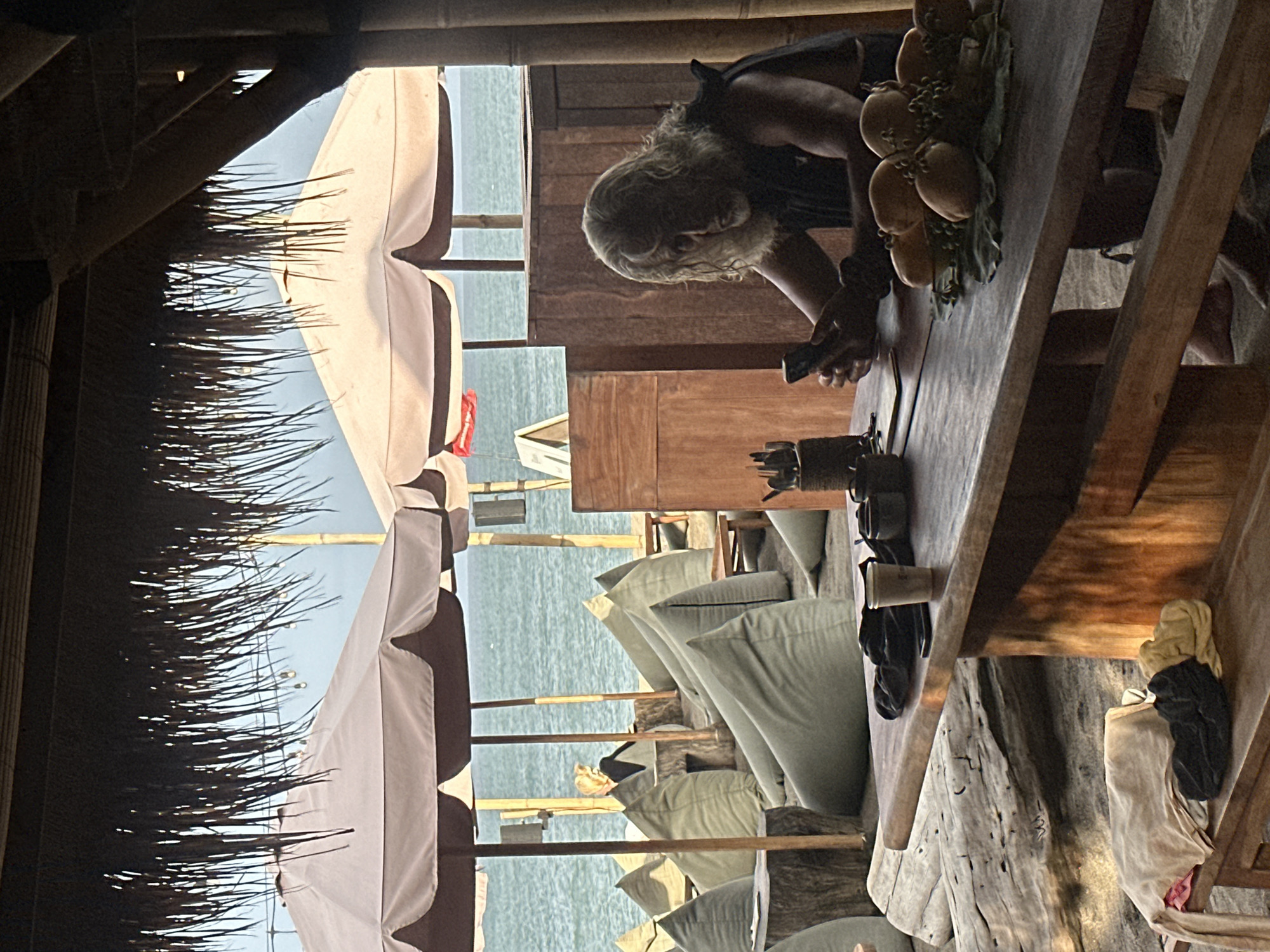 This image depicts a dimly lit, tropical setting, likely a beachside establishment in North Kuta, Indonesia. The perspective is from underneath a thatched roof structure, looking out towards a body of water, possibly the ocean.
The main subject is a person with long, light-colored hair, hunched over a wooden table and looking at a smartphone. They are wearing a dark, possibly sleeveless top. On the table, in front of the person, is a decorative arrangement of what appear to be artificial fruits, possibly mangoes and grapes. There are also various objects on the table, including what seem to be small cups or containers.
The scene is characterized by natural materials, with prominent use of wood and thatch. Several large, cream-colored umbrellas with dark trim are visible in the background, suggesting an outdoor seating area. Beyond the umbrellas, the calm, turquoise water of the sea is visible under a bright sky. The lighting suggests it is daytime, likely late afternoon or early evening due to the warm, golden tones and soft shadows.
In the foreground, the texture of the thatched roof is evident, with long strands of dried grass casting shadows. The overall atmosphere feels relaxed and somewhat rustic. Some items of clothing are draped over a wooden beam in the lower right portion of the image. No text is visible in the image.