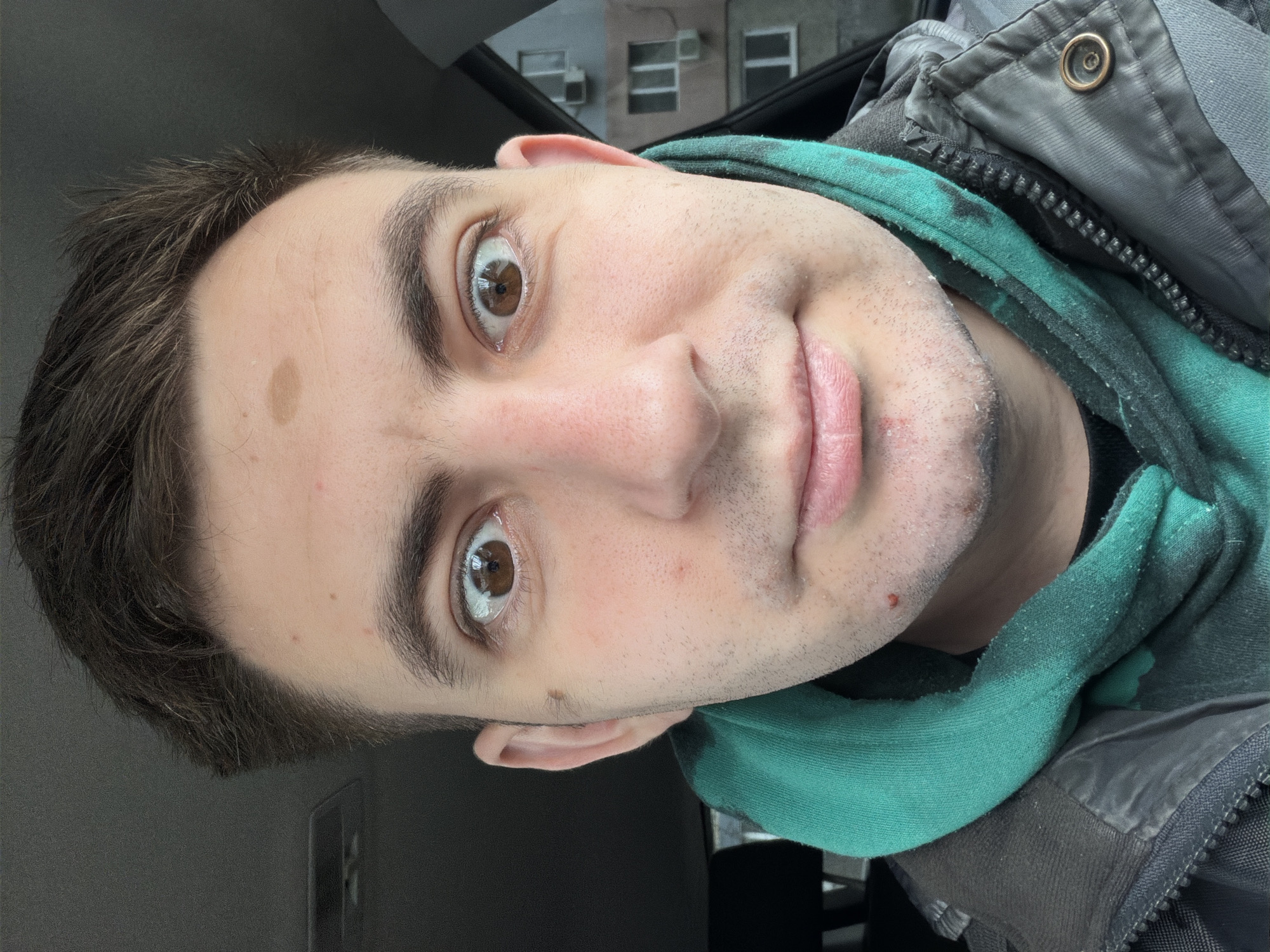 An off-center, close-up selfie depicts a fair-skinned male with short dark brown hair and brown eyes. He has light stubble and a mole on his forehead. He is wearing a dark grey jacket with a metal button and a green hooded sweatshirt underneath. The photo appears to be taken indoors, possibly in a car, with a blurred exterior background showing an indistinct building. The location in Varna, Bulgaria cannot be inferred from the image.