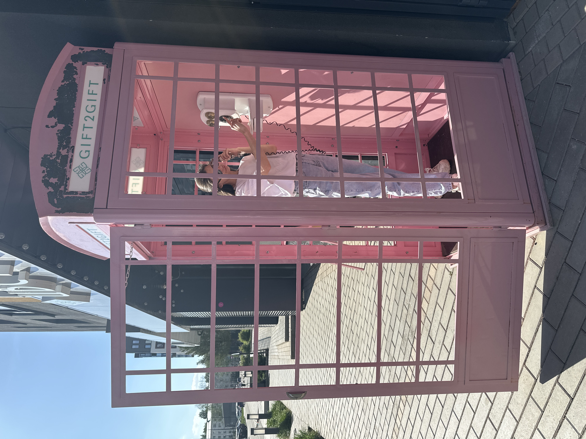 A pink telephone booth stands on a paved surface in an urban setting in Izhevsk, Russia. The booth has an open door and a person inside, holding a white telephone receiver to their ear. The booth is adorned with the text "GIFT2GIFT" on the top. The person inside is wearing a white t-shirt and blue jeans. Outside the booth, there are buildings, greenery, and street lighting.