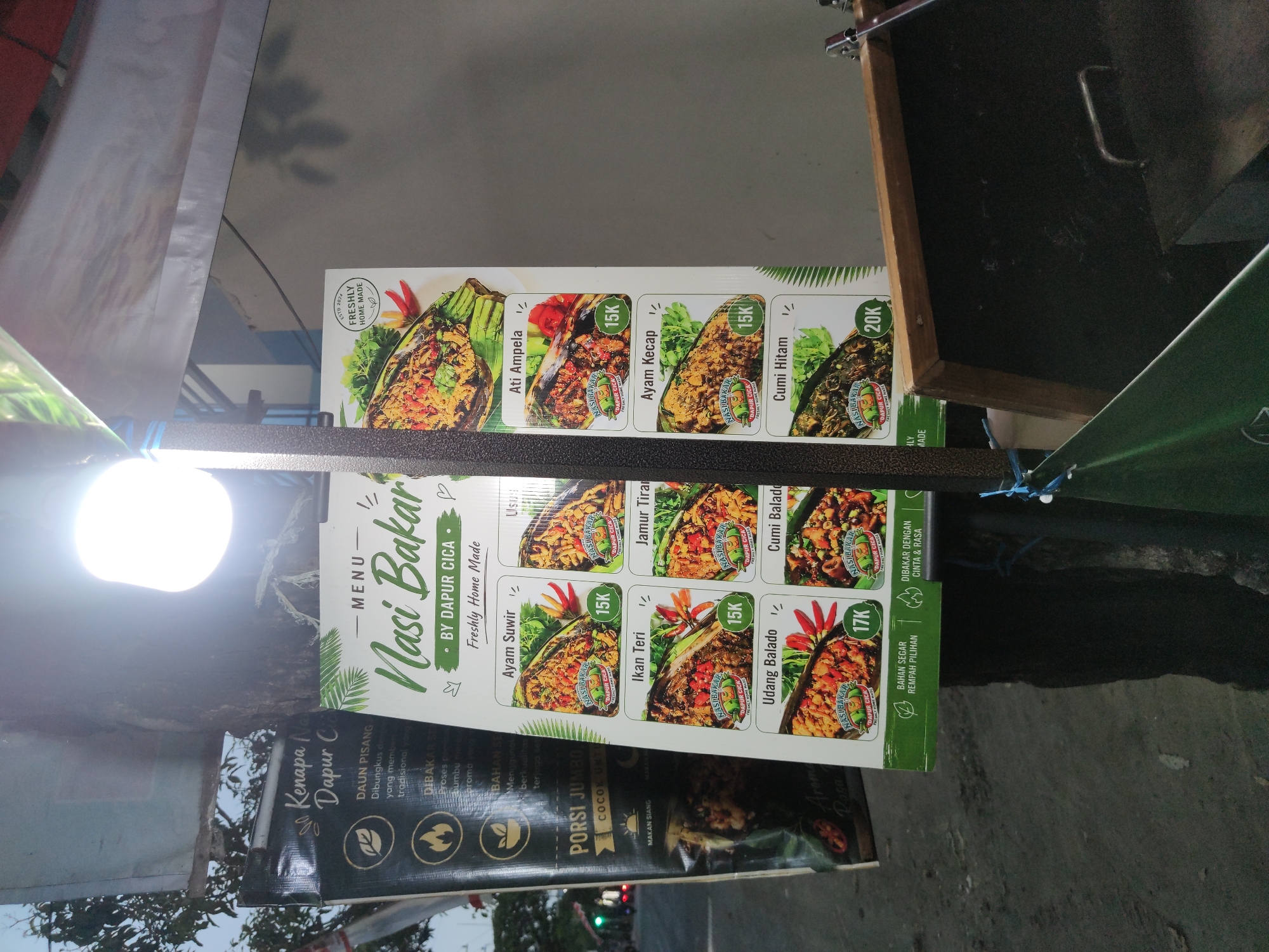 The image shows a close-up of a food menu for "Nasi Bakar" (grilled rice) from "Dapur Cica" (Cica's Kitchen). The menu is displayed on a white board with green accents and features pictures of various dishes with their prices. The menu items include "Ayam Suwir" (shredded chicken), "Ikan Teri" (anchovies), "Udang Balado" (shrimp with chili paste), "Cumi Hitam" (black squid), "Ayam Kecap" (soy sauce chicken), and "Jamur Tiram" (oyster mushroom). Prices range from 15K to 20K. To the left of the menu, a dark green banner with white text promotes "Kenapa Nasi Bakar" (Why Nasi Bakar), highlighting features like "Daun Pisang" (banana leaf wrapping), "Dibakar" (grilled), and "Bahan Segar" (fresh ingredients). The menu and banner are attached to a tree, suggesting an outdoor food stall or eatery. A bright light bulb illuminates the scene from above, casting shadows. In the background, blurred street lights and trees suggest an urban environment at dusk or night. The overall impression is that of a casual, street-side food vendor in Surabaya, Indonesia, likely operating in the evening.