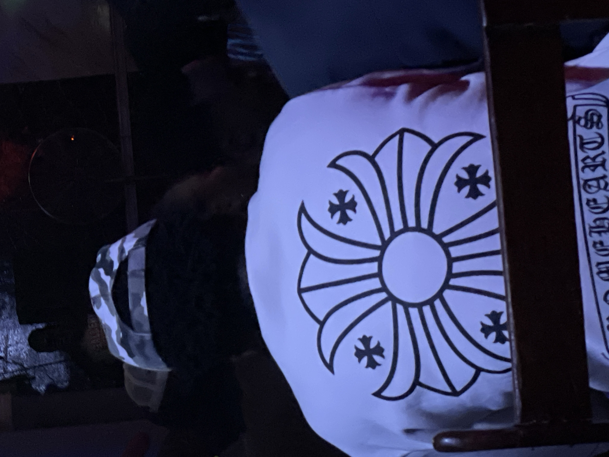 The image shows the back of a person wearing a white long-sleeved shirt with a large, stylized floral design in black on the back. The design features a central circle with radiating petal-like shapes, each containing a smaller cross symbol. The person is also wearing a camouflage baseball cap. The setting appears to be dimly lit, possibly an indoor venue. The location is not specified in the image or provided information as being in Durumi, Nigeria.