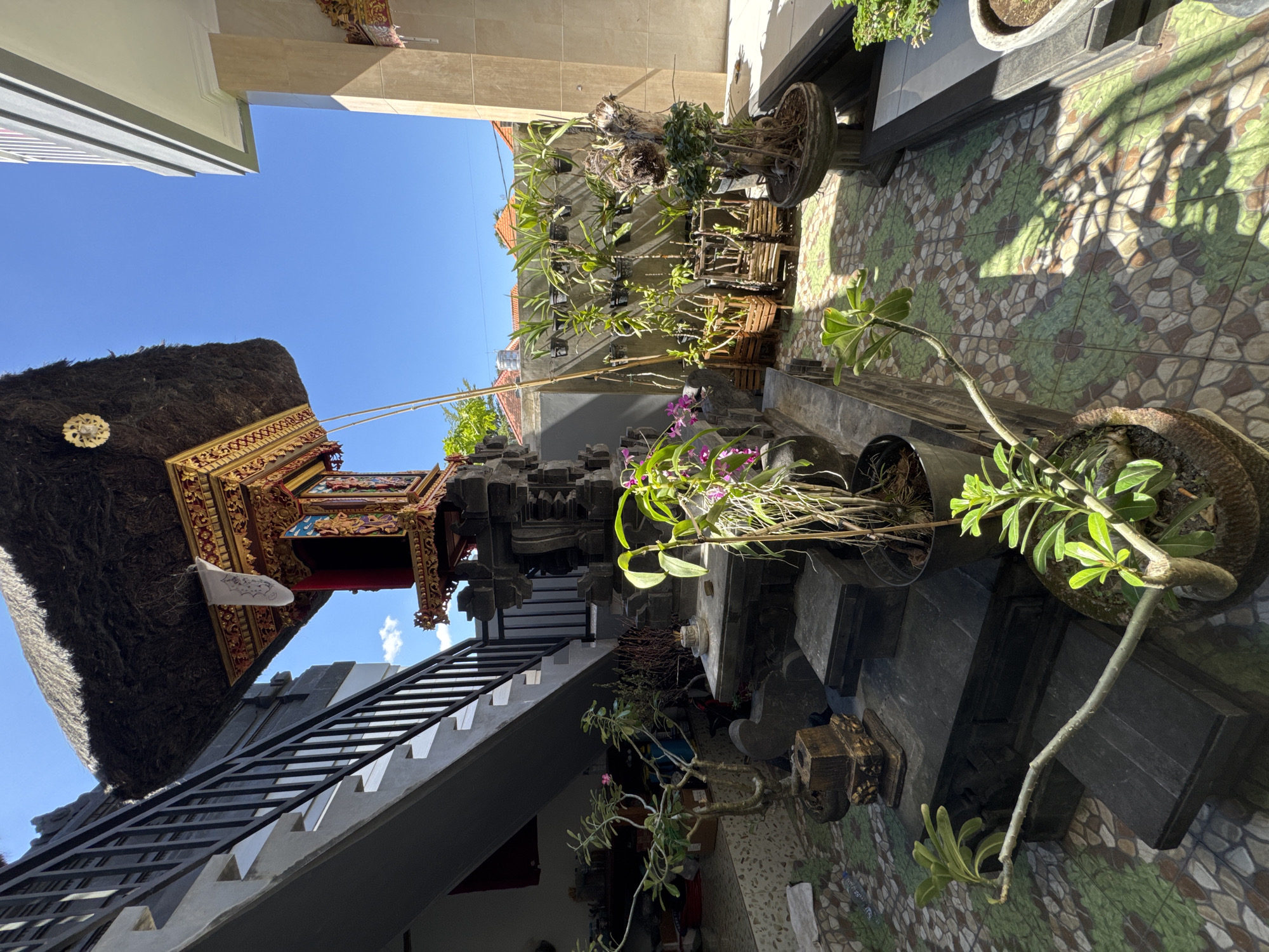 The image depicts a scene in Kuta, Indonesia, showcasing traditional Balinese architecture and landscaping. On the left, a building features a thatched roof and intricate carvings. A set of stairs with a black metal railing leads upwards. Various plants and pots are situated on the right, adding greenery to the space. The ground is paved with decorative tiles in a pattern of green, brown, and tan. The sky is clear and blue, indicating a sunny day. There are no people visible in the frame.