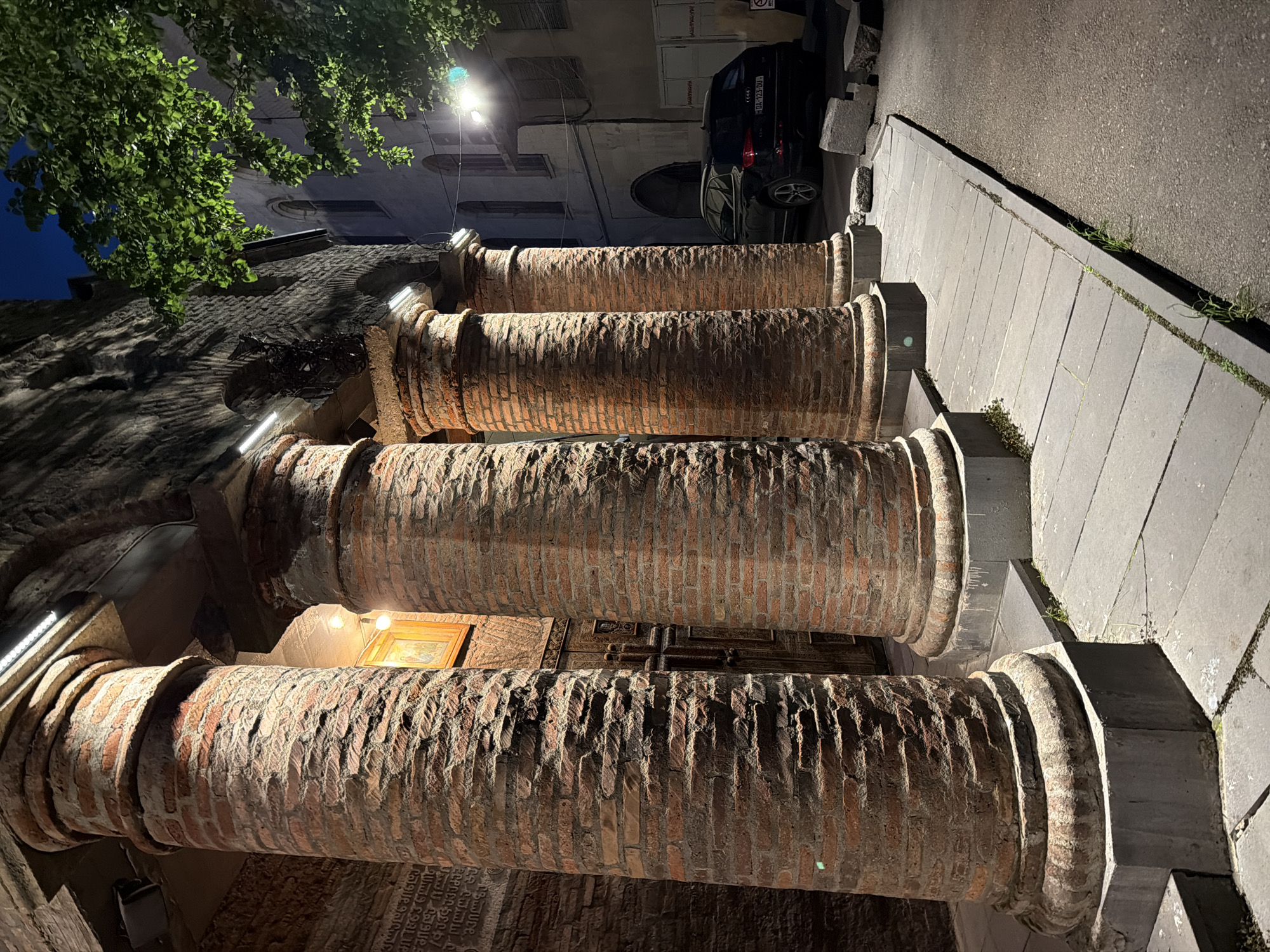 Four brick columns line a stone staircase leading to a dark entrance. The columns are topped with gray stone capitals. Behind the columns, carved wooden doors are visible, along with a framed picture on the wall. To the right of the staircase is a gray stone walkway leading to a dark asphalt street where a black car is parked. Overhead, a street light illuminates the scene, while tree branches with green leaves are visible in the upper left. The image appears to be taken at night. The location is likely in Tbilisi, Georgia.