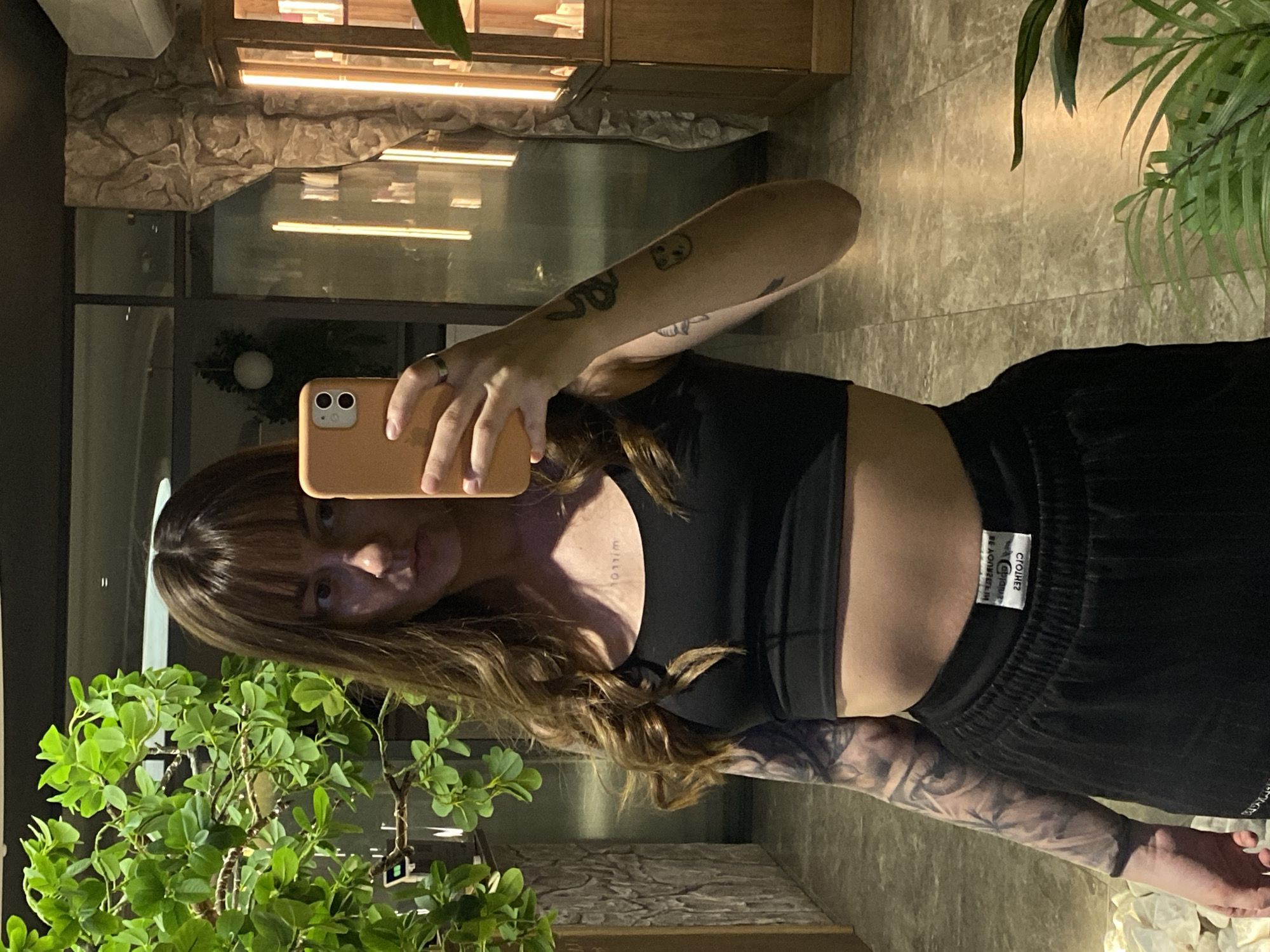 A woman is taking a selfie with her smartphone, which is pale orange, in an indoor location in what may be Votkinsk, Russia. She is wearing a black crop top and black sweatpants with a white tag visible on the waistband. The woman has light brown hair with bangs. Her left arm has a visible tattoo. The background includes indoor plants, stone textured walls, and a display cabinet.