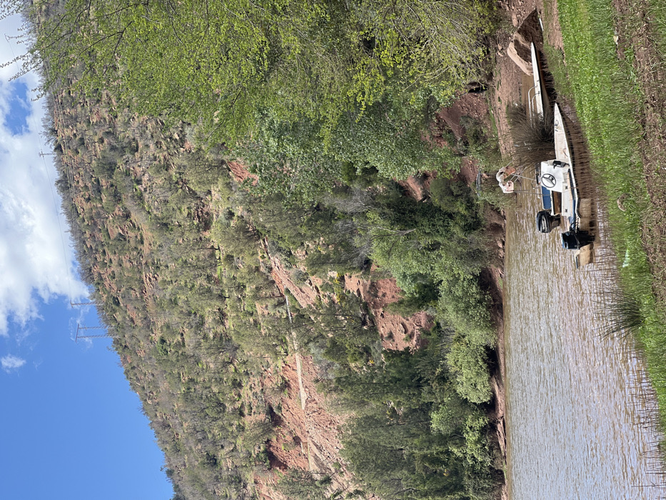 The scene, located in Ait Ouaarda, Morocco, depicts a body of brownish-green water meeting a vegetated and earthen bank. In the right foreground, two motorboats are partially beached: a larger, light-colored vessel equipped with a steering console and a black outboard motor, and a smaller, darker boat with a second black outboard motor positioned behind it. A third, smaller, light-colored flat-bottomed vessel is pulled further onto the immediate shore. A white, flamingo-shaped decorative object stands near the larger boat. The bank is covered with green grass and reeds in the immediate foreground, transitioning to denser green trees and shrubs further up the slope. A steep hillside, characterized by exposed reddish-brown soil and rocks, rises prominently in the background. Sparse to moderate vegetation, primarily small trees and scrub with green foliage, covers the hillside. A narrow, light-colored dirt track is visible traversing the mid-slope of the hill. Overhead, multiple metal electricity pylons with associated power lines are positioned along the hilltop, silhouetted against a clear blue sky featuring scattered white cumulus clouds. The scene is illuminated by natural daylight. No individuals are clearly discernible engaging in specific actions or interactions.