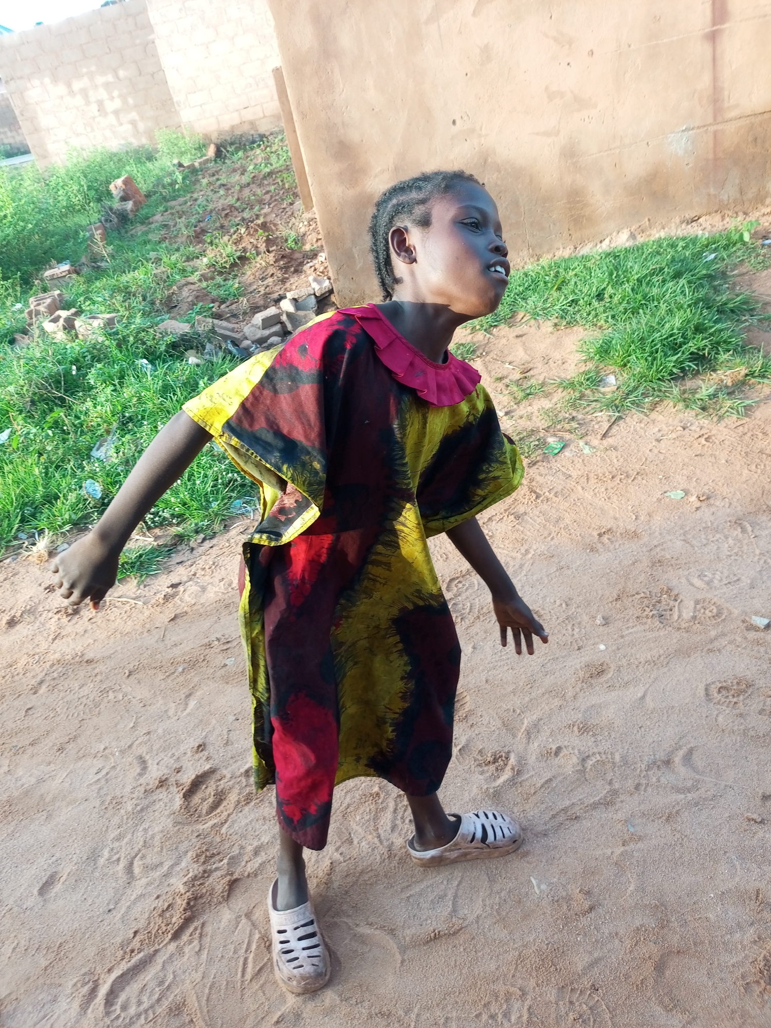 A young African child stands in a sandy area in front of a tan-colored building. The child is wearing a colorful, loose-fitting dress with red, yellow, and black patterns. The dress has a red ruffled collar and short, wide sleeves. The child has braided hair and is looking upwards and to the right with an open mouth. They are wearing white sandals with cut-out details. The ground is sandy with some footprints visible. Behind the child, there is a partially constructed building with light-colored bricks and a tan wall. Green grass and some scattered bricks are visible in the background, suggesting an outdoor environment, possibly in a semi-developed area of Tempure, Nigeria.