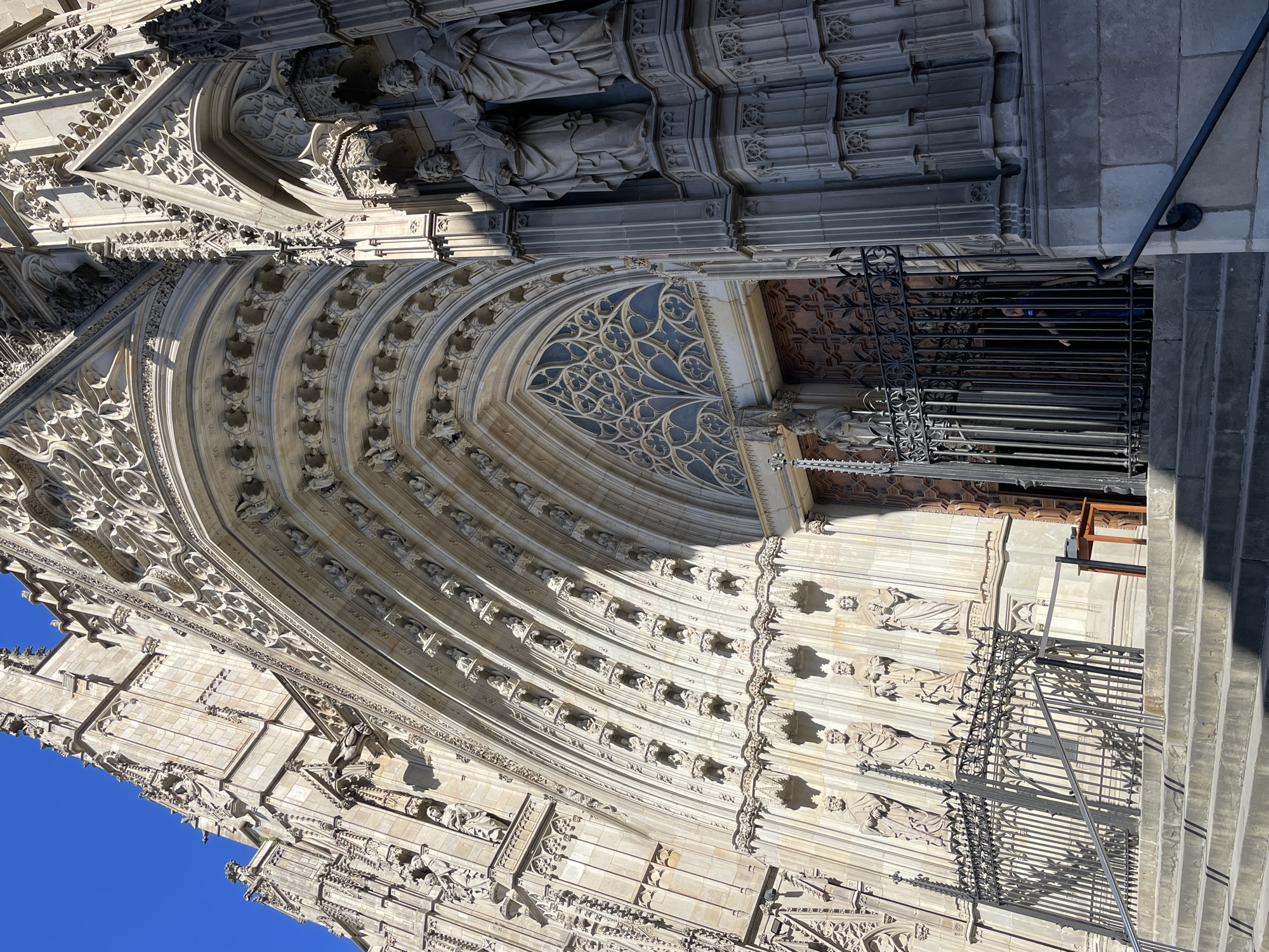 The image shows a close-up, low-angle shot of the facade of a large, ornate building, likely a cathedral or significant historical structure, under a clear blue sky. The building's architecture is characterized by intricate stone carvings and details, predominantly in a light tan or beige color. Arched doorways and windows, embellished with decorative patterns, are prominent features. Statues are integrated into the facade. A metal gate or fence is visible in front of a doorway, and a staircase is present on the right side of the frame. The image is not set in Barcelona, Spain, as the architecture does not reflect the distinctive style typically associated with the city. Instead, the building in the picture is Seville Cathedral.