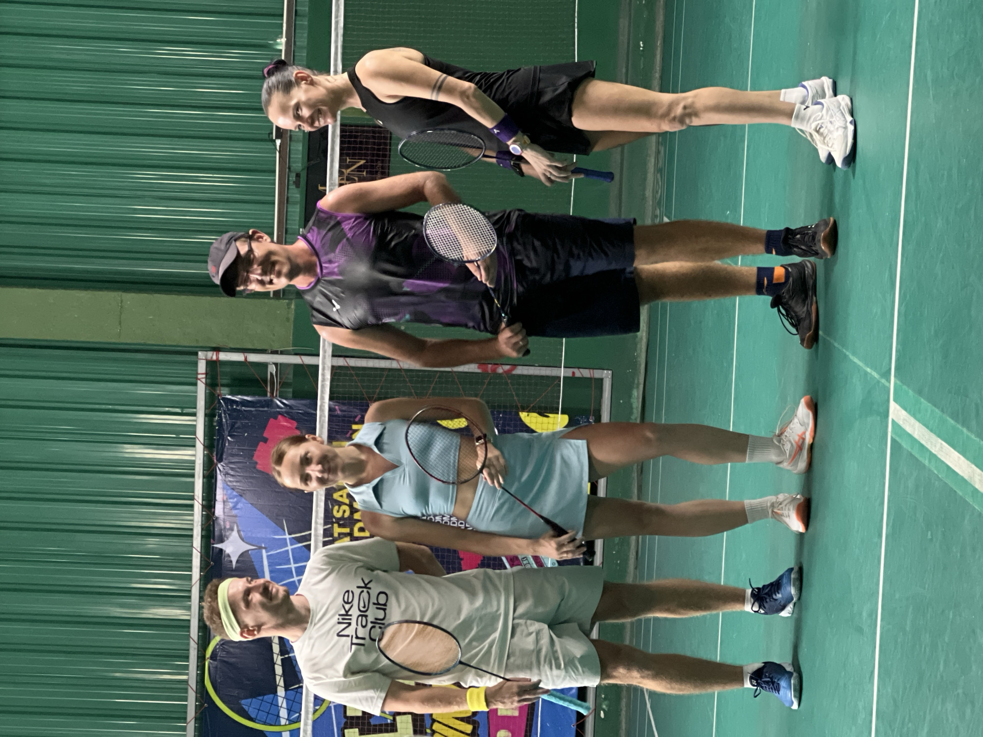 Four individuals, two men and two women, stand on a badminton court in a sports facility. The court is green with white lines. The background consists of a green ribbed wall and a banner with text and graphics. All four people are holding badminton rackets and appear to be posing for a photograph. They are dressed in athletic wear. The man on the far left wears a white Nike t-shirt and light-colored shorts. The woman next to him is in a light blue athletic set consisting of a crop top and skirt. The second man from the left is wearing a dark sleeveless shirt with purple accents and dark shorts. The woman on the far right is in a black sleeveless top and black skirt. The lighting suggests an indoor environment, possibly an evening or dimly lit facility. The overall scene depicts a group ready to play or having just finished playing badminton. The location is identified as a badminton court in Baan Bang Rak, Thailand.