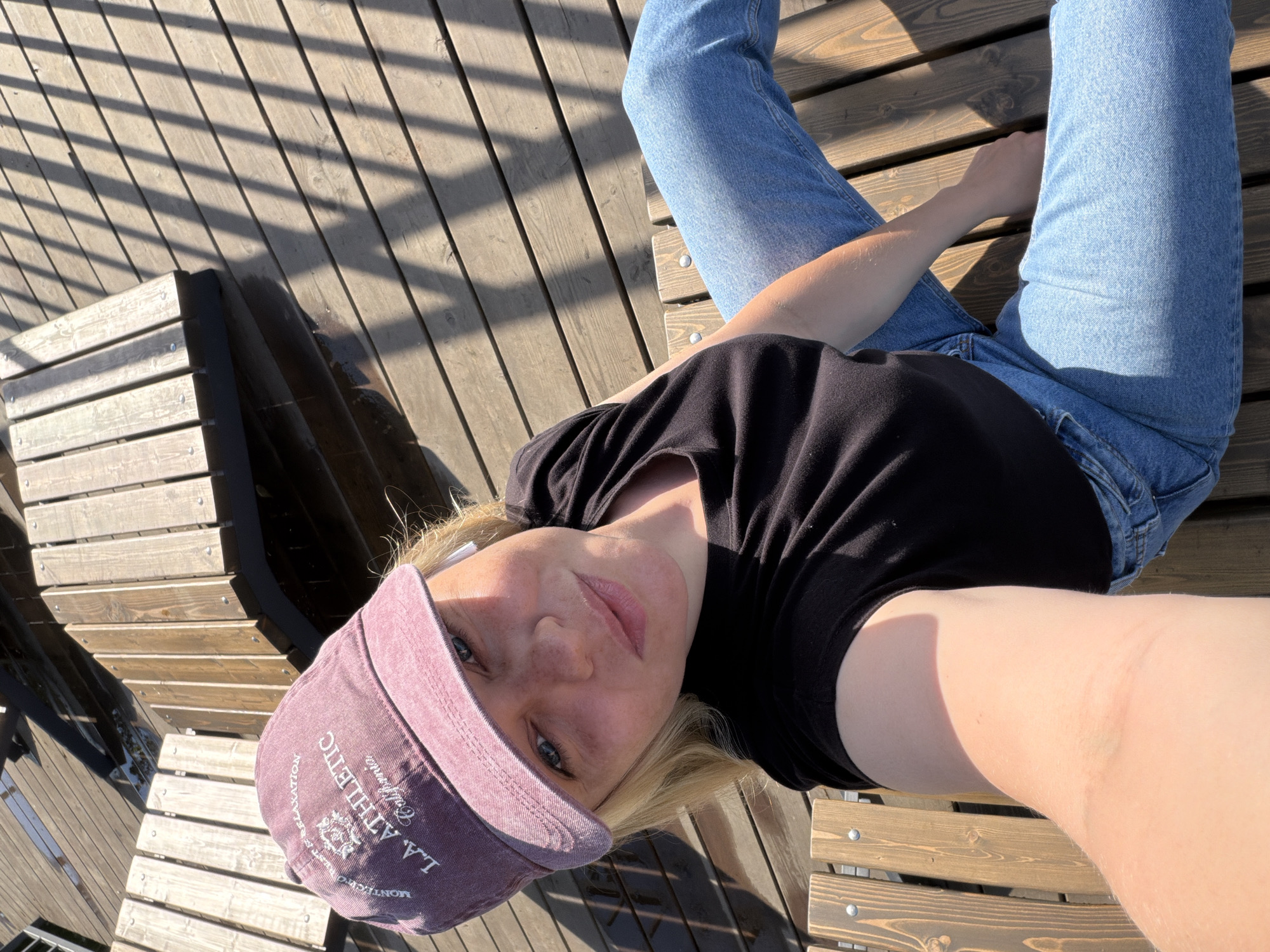 A person wearing blue jeans, a black t-shirt, and a purple "L.A. Athletic" baseball cap is sitting on a wooden bench. The person is holding the camera and taking a selfie. The location appears to be an outdoor public space with wooden benches and decking. There is a city park in Votkinsk, Russia.