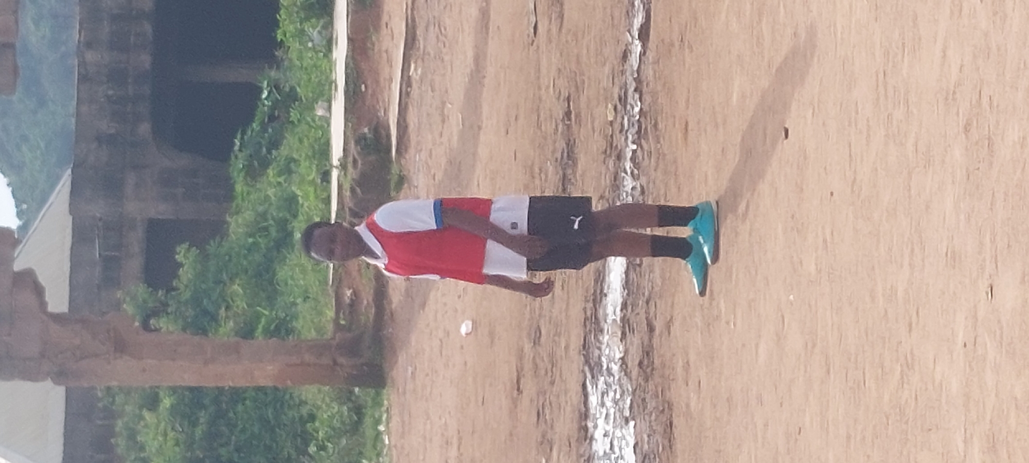 A young person stands in a dirt field in Bukuru, Nigeria, wearing a white shirt with a red vest over it and black shorts. The person is also wearing dark socks and bright blue athletic shoes. The field is uneven with patches of dried mud and some debris. In the background, there is a large, weathered stone structure with arched openings and some overgrown vegetation. The sky is overcast.