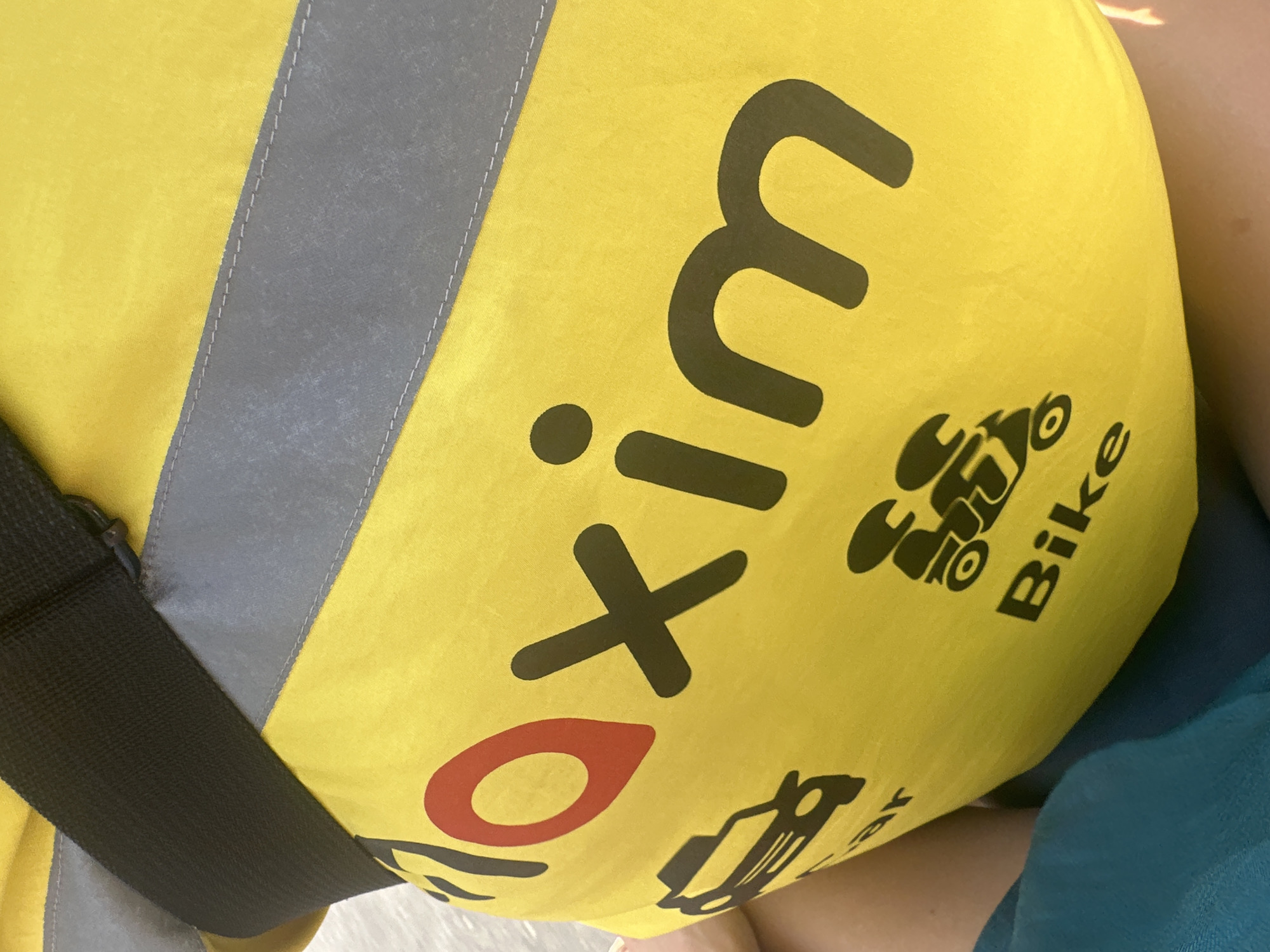 The image shows a close-up of a yellow delivery bag with black and red text and graphics, likely belonging to a delivery service. The text "maxim" is prominently displayed in large black letters, with a red location pin symbol integrated into the letter 'o'. Below the word "maxim", there are icons representing a car and two motorcycles, with the word "Bike" beneath the motorcycles. The bag appears to be worn by a person, with a black strap visible at the top and a portion of the person's legs covered by a blue fabric. The overall scene suggests a commercial delivery context. The provided location of Bac Nha Trang Ward, Vietnam, is context provided to the model, not visually evident in the image.