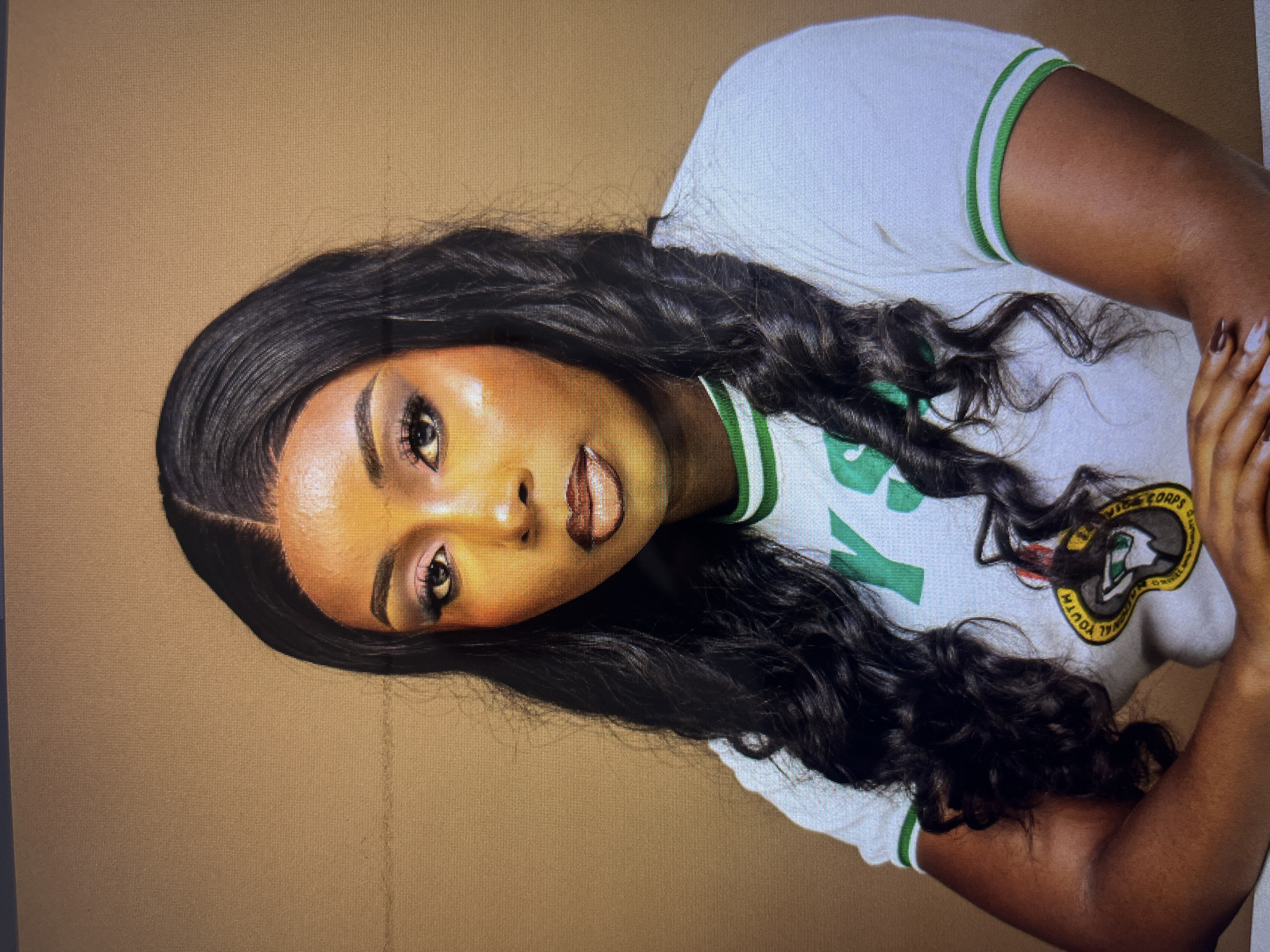 A studio portrait of a young Black woman with long, dark, wavy hair. She is wearing a white t-shirt with green and white stripes on the collar and sleeves. The t-shirt has a green logo on the front. She is wearing makeup, including eyeshadow and lipstick. Her arms are crossed in front of her. The background is a solid, light brown color. The lighting is bright, highlighting her face and shoulders. The image appears to be a professionally taken photograph, likely in a studio setting. There are no visible environmental or architectural clues to indicate a specific location within Gura Top, Nigeria.