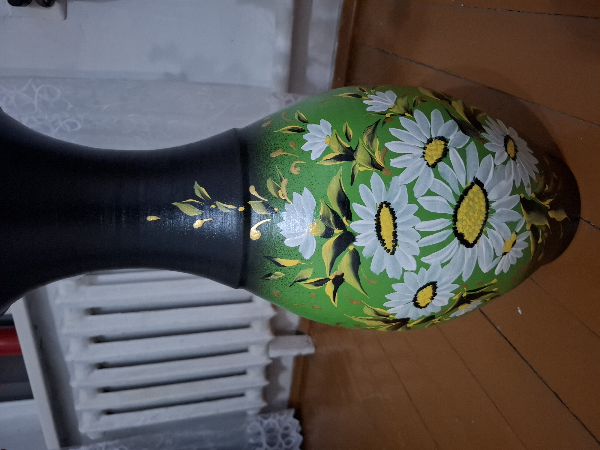 A hand-painted ceramic vase with a black top section and a green body adorned with white daisies and yellow-green leaves is displayed on a wooden floor indoors. In the background, a white radiator and a white lace curtain are visible. The overall scene is lit by artificial lighting.