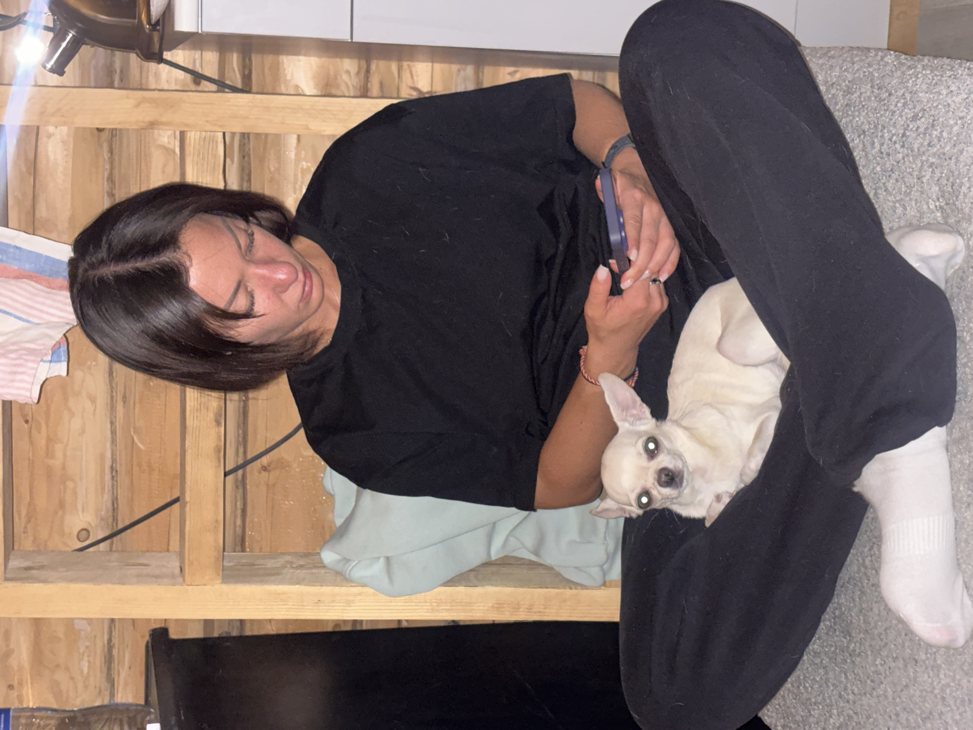 A woman sits on a wooden structure, possibly a makeshift platform or elevated seating area. She is wearing a black t-shirt and black sweatpants. She is holding a smartphone and appears to be looking at it. A small, cream-colored dog, possibly a Chihuahua, is nestled on her lap. The woman is wearing white socks. The background includes a wooden wall, and parts of what looks like a light-colored blanket or cloth are visible. The environment suggests an indoor location that is rustic or unfinished, with the presence of wood and the lighting. There is no direct visual information available in the image to connect it to the city of Emel'yanovo, Russia.