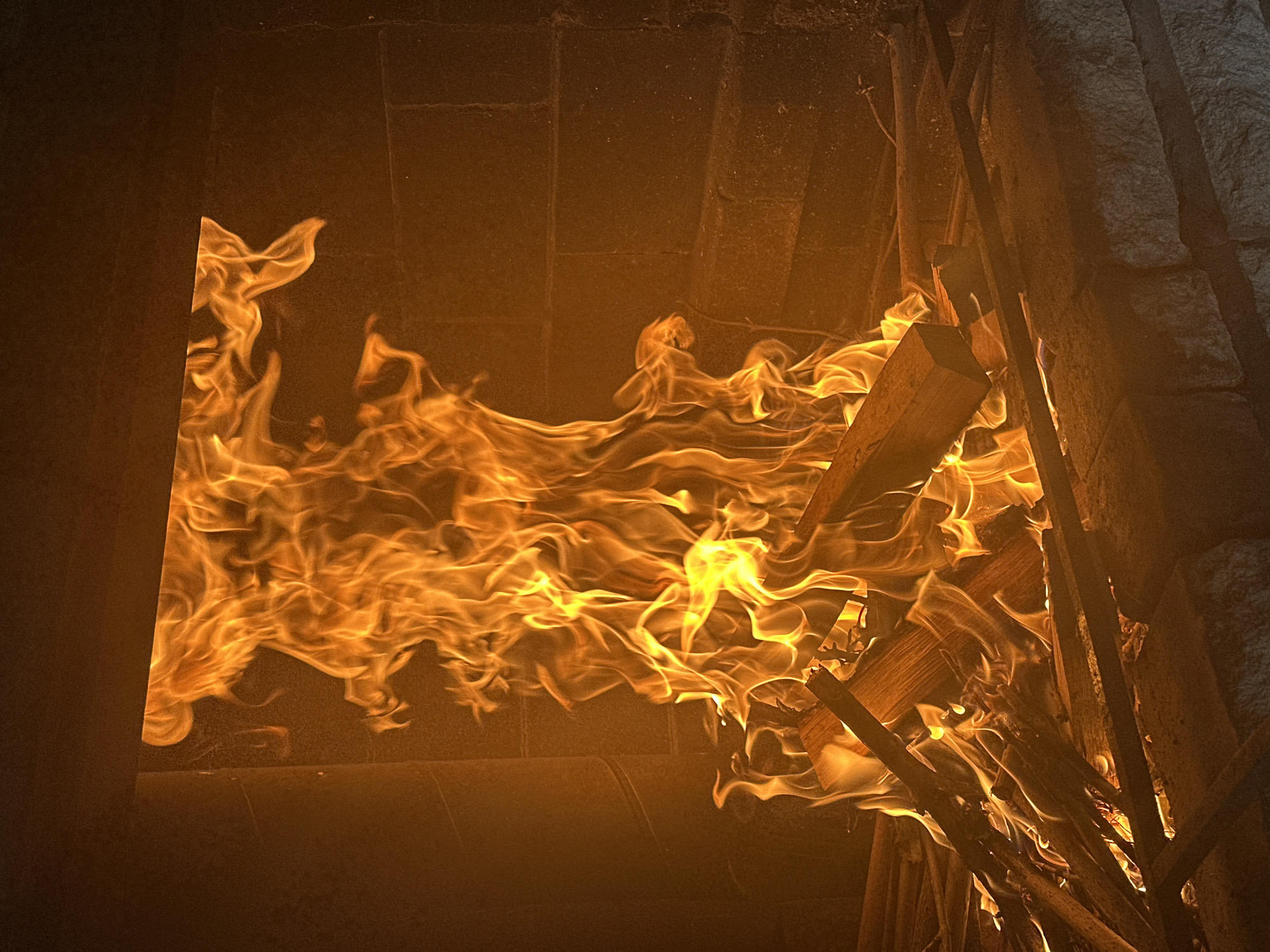 The image depicts a fire burning inside a brick-lined fireplace or enclosure. Flames are actively consuming wooden materials. The structure appears to be constructed from dark-colored bricks or stones. The setting is not identifiable as being in Aviator, Russia from the visual information available.