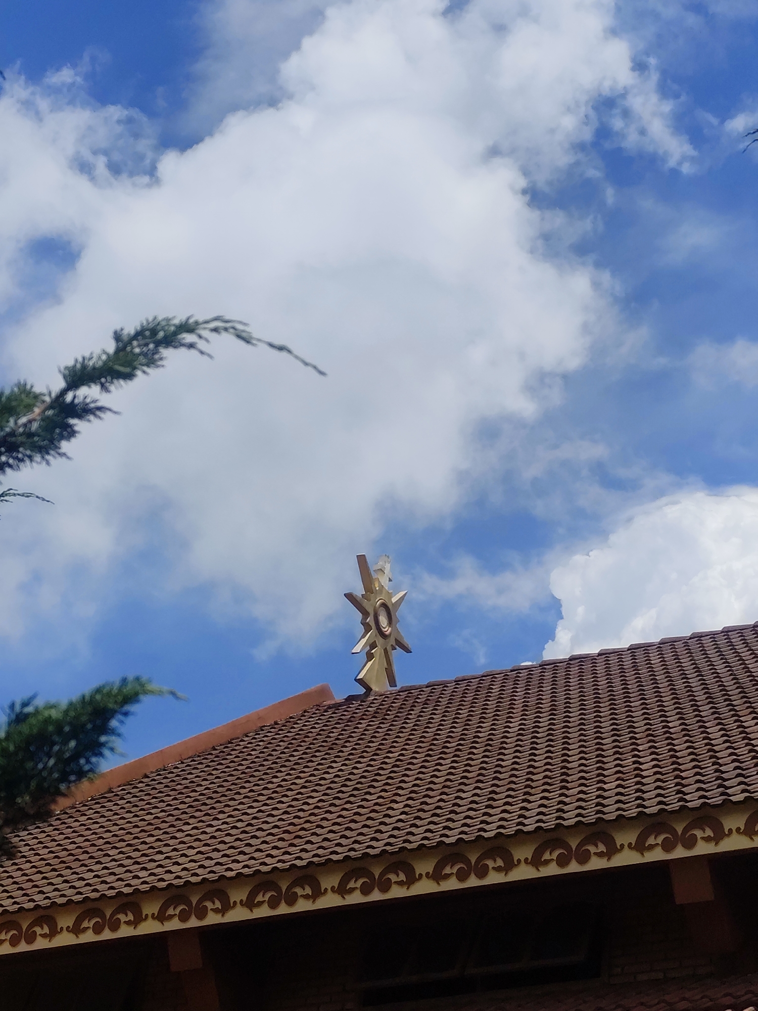 The image shows the roof of a building against a blue sky with white clouds. A distinctive, golden, star-shaped ornament with a circular center is positioned at the peak of the tiled roof. The roof tiles are a reddish-brown color and have a wavy pattern. Decorative trim with a scrolled design runs along the edge of the roof. To the left, a branch of a dark green evergreen tree is visible, arching over the roof. The scene appears to be outdoors, and the architectural style of the roof and ornament suggests it could be a religious building or a traditional structure in Da Lat, Vietnam.