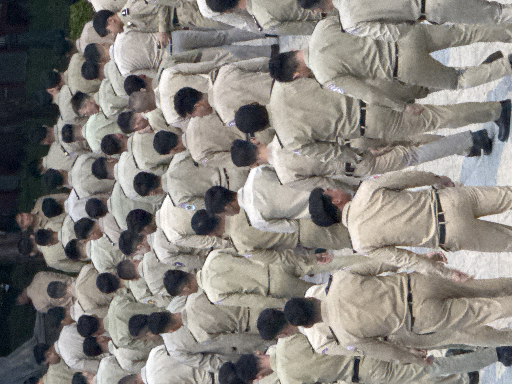 In Da Lat, Vietnam, a large group of young men, likely students or recruits, are gathered in what appears to be an outdoor educational or ceremonial setting. They are all wearing matching tan uniforms consisting of long-sleeved shirts and trousers, with black belts and dark footwear. Many of the shirts have epaulets and some have small crests or patches on the sleeves. The men are mostly facing away from the camera, with a few individuals looking to the side or towards each other. Their stances suggest they are either waiting, marching, or participating in a structured activity. The background is mostly out of focus, but hints of trees and foliage suggest a park-like or campus environment. The overall impression is one of order and uniformity within a youthful demographic.