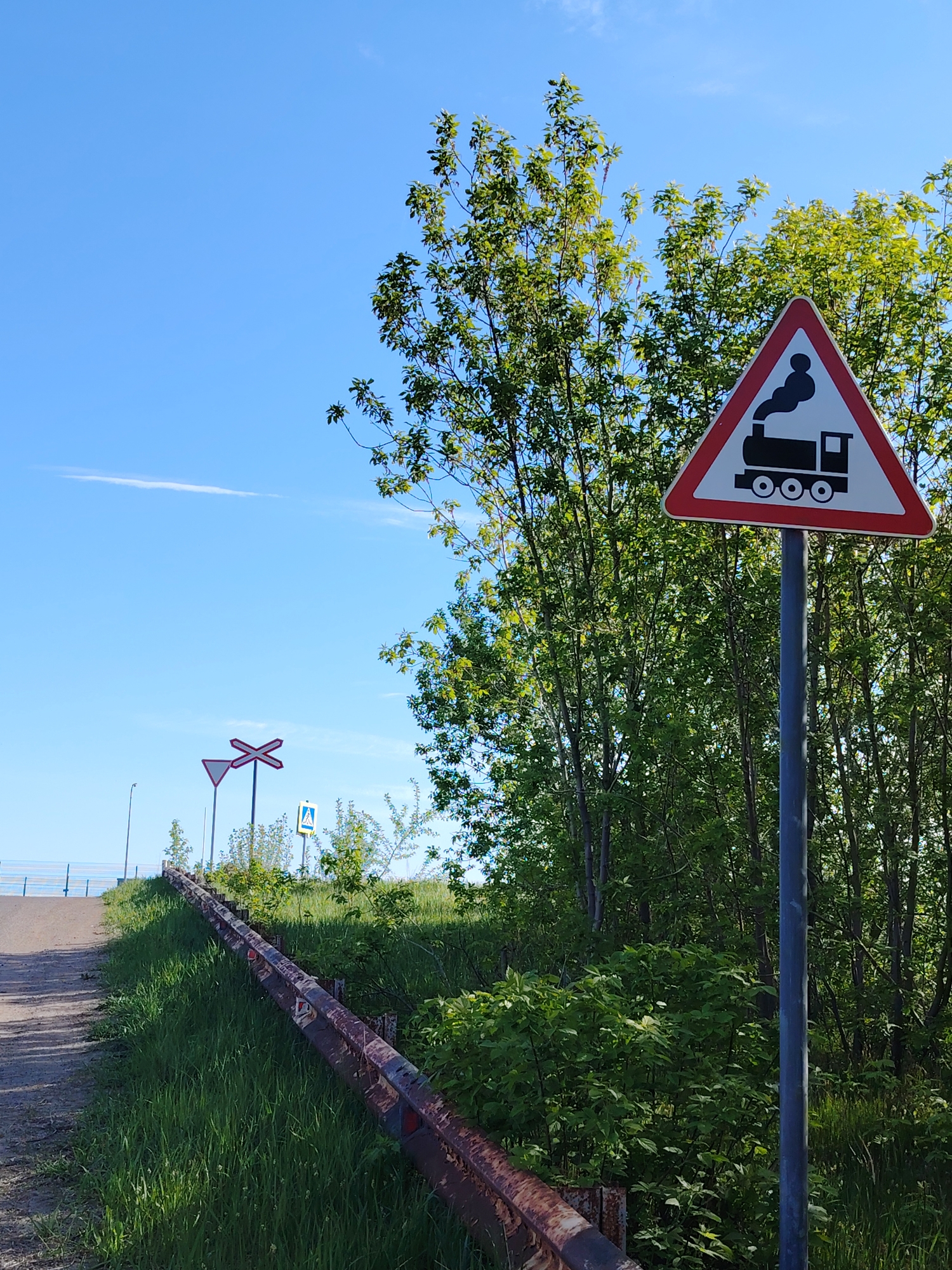 A sunny, outdoor scene depicts a road leading uphill, flanked by a rusty guardrail and greenery. In the background, other road signs are visible, along with a possible body of water in the distance. In the foreground, a triangular traffic sign with a red border and an image of a steam train stands prominently. The foliage surrounding the sign appears lush and full. The image does not contain information about the location in the city of Chaikovsky, Russia.