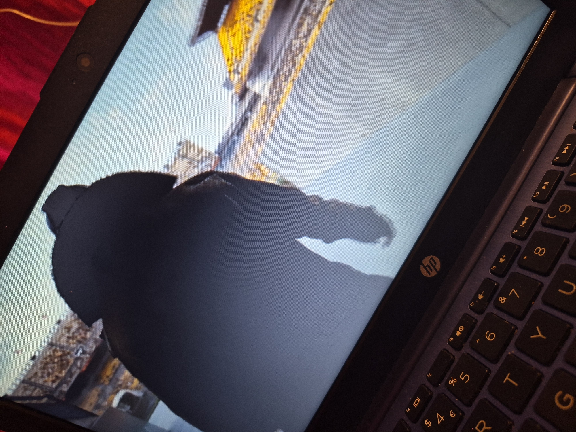 The image shows a laptop screen displaying a video or game. On the screen, a person is seen from behind, walking away from the camera. They are wearing a dark hooded jacket. The background appears to be a stadium with yellow seating and structures. In the foreground, part of the laptop's keyboard is visible, showing keys with symbols and letters, including the HP logo. The laptop is positioned on a dark surface, possibly a table or desk. The red fabric in the upper right corner suggests the environment surrounding the laptop. There is no information in the image to suggest the location is in Sandwell, United Kingdom.