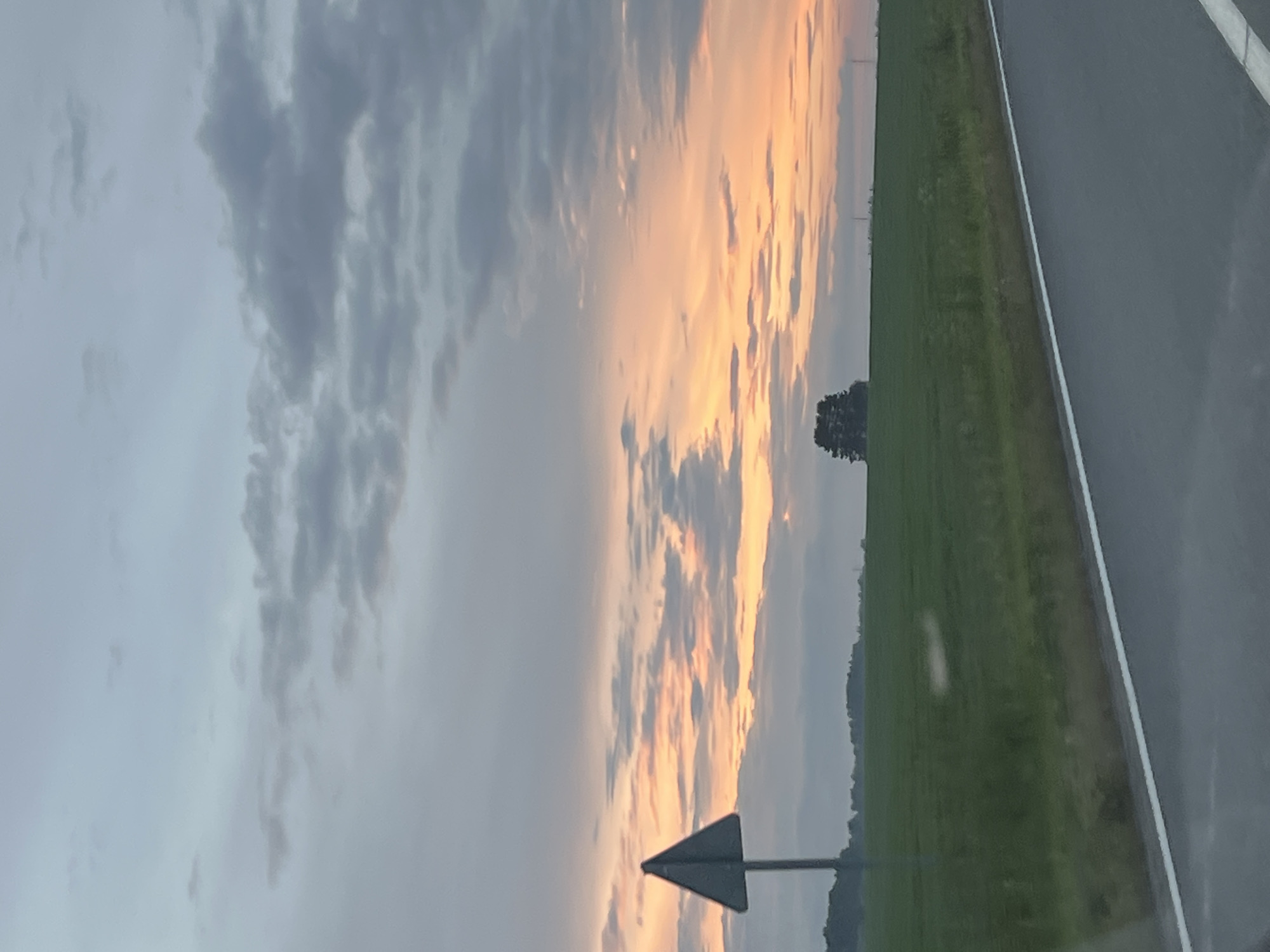 This image depicts a rural roadside scene in Поварёнки, Russia, at dusk. The sky is filled with dramatic clouds, tinged with the warm hues of sunset, casting an orange glow on some of the clouds while others remain a soft grey. A single, dark tree stands silhouetted against the colorful sky in the distance.
To the right of the image, a paved road runs parallel to a vast, green field. The field is likely agricultural, with subtle variations in texture suggesting crops. A white line marks the edge of the road, bordering it from the field. A dark triangular sign on a pole is visible in the lower left portion of the image, pointing to the right, indicating a road direction or hazard. The overall atmosphere suggests a peaceful, natural setting as daylight fades. The weather appears to be clear with some cloud cover, typical for the evening. No people are visible in the image.