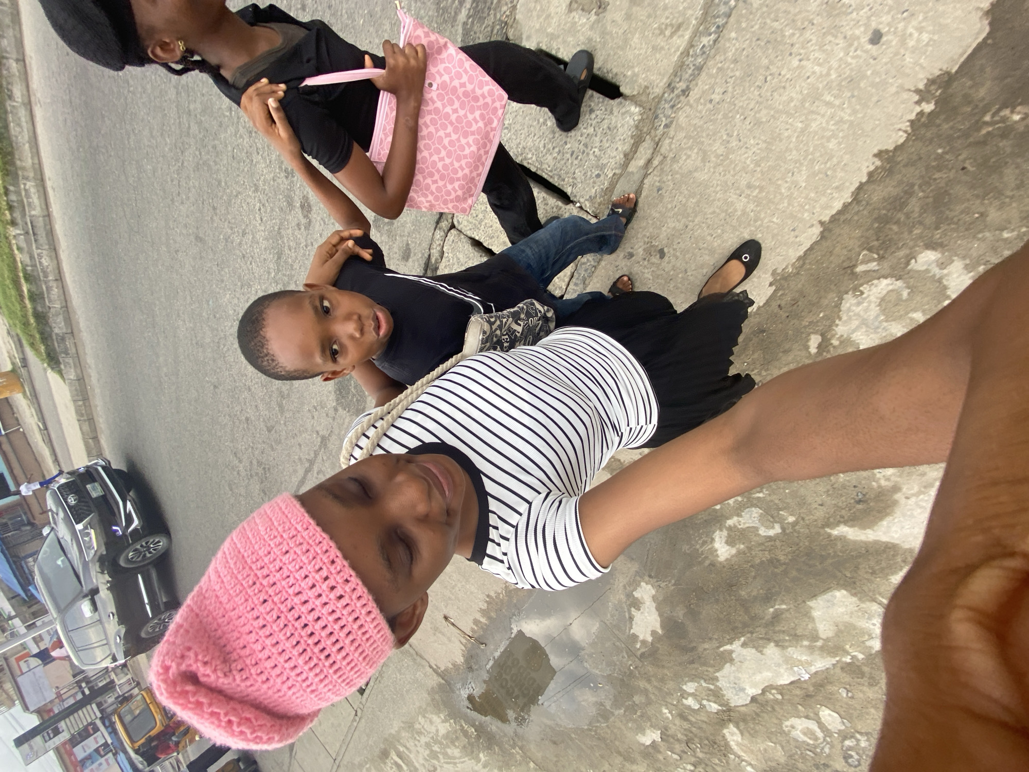 Three children stand on a sidewalk in Unknown, Unknown. One is wearing a pink knit cap and a black and white striped shirt, another is wearing a black t-shirt and jeans, and the third is wearing a black shirt, pants, and a black cap and holding a pink purse. There is a puddle of water on the sidewalk. A car and other vehicles are visible in the background.
