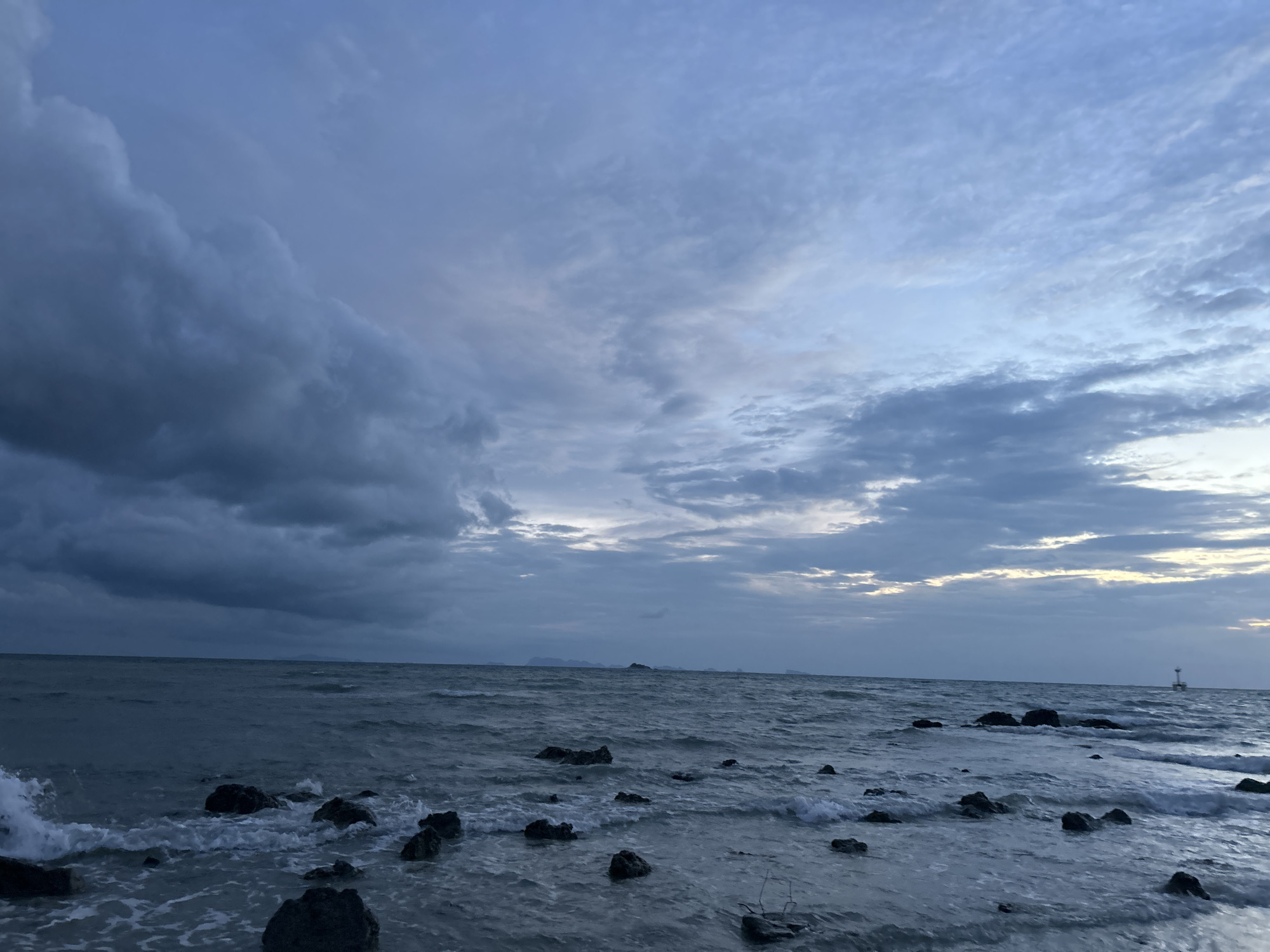 The image depicts a body of water with waves breaking against scattered rocks in the foreground. The sky above is filled with dramatic, dark clouds, suggesting an overcast or stormy day. In the far distance, a faint outline of land can be seen, and a small lighthouse or marker stands on the horizon. The overall impression is a moody and natural scene, likely a coastal area.