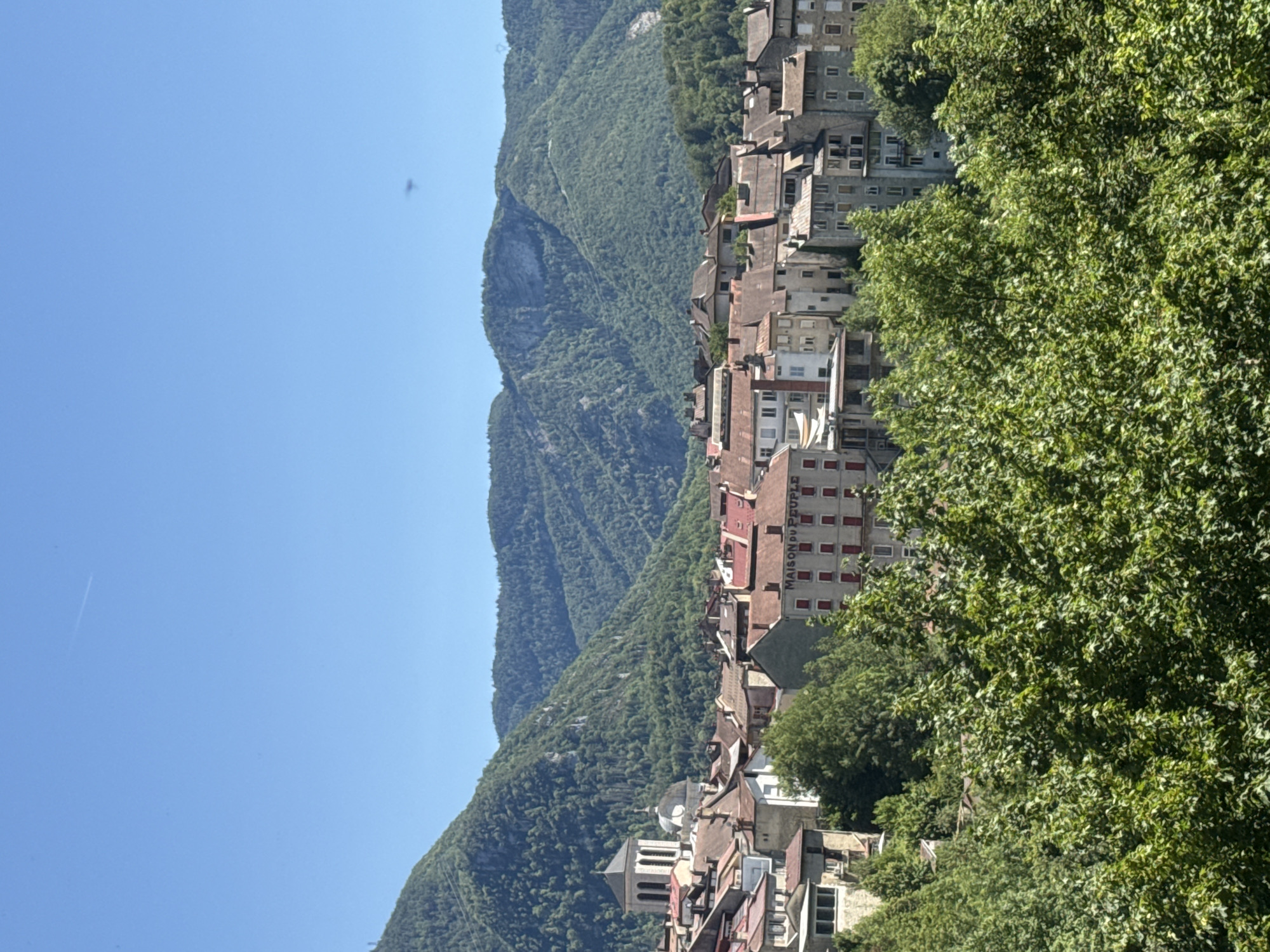 The image shows a view of the city of Saint-Claude, France, nestled in a valley surrounded by lush green mountains. The city features a dense collection of buildings with terracotta roofs, characteristic of French architecture. Prominent structures include a church with a tall bell tower and a dome, as well as buildings with visible signs, one of which reads "MAISON DU PEUPLE" (People's House). The foreground is dominated by the dense foliage of green trees. The sky is a clear, vibrant blue. There are no people visible in the image. The overall scene depicts a picturesque and tranquil European town.