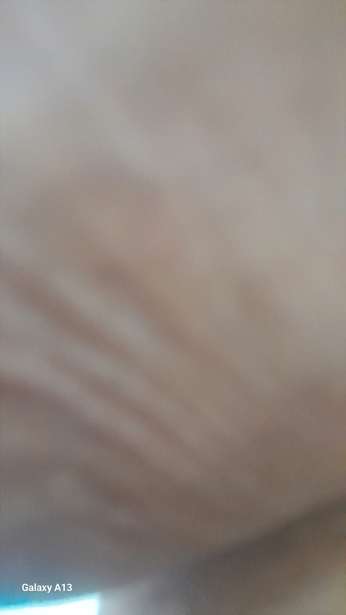 The image is a close-up, out-of-focus shot of a beige surface with subtle, diagonal lines or ridges visible. The texture appears somewhat soft or fabric-like. In the bottom left corner, the text "Galaxy A13" is visible, suggesting this is a screenshot or a photo taken with a Samsung Galaxy A13 phone. The image is entirely devoid of any contextual information related to the city of Hadipolo, Indonesia, or any discernible subjects, objects, actions, or interactions. The overall impression is of a very abstract and blurry visual.