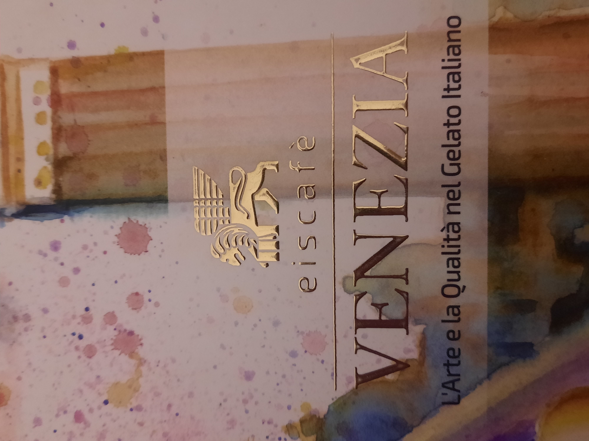 The image shows a close-up of a sign or menu featuring a golden winged lion emblem above the text "eiscafè VENEZIA L'Arte e la Qualità nel Gelato Italiano". The background is a light-colored surface with watercolor splatters in pink, purple, and yellow. The overall aesthetic suggests a high-end Italian ice cream shop. There is no information in the image to indicate a location in Rosenheim, Germany.