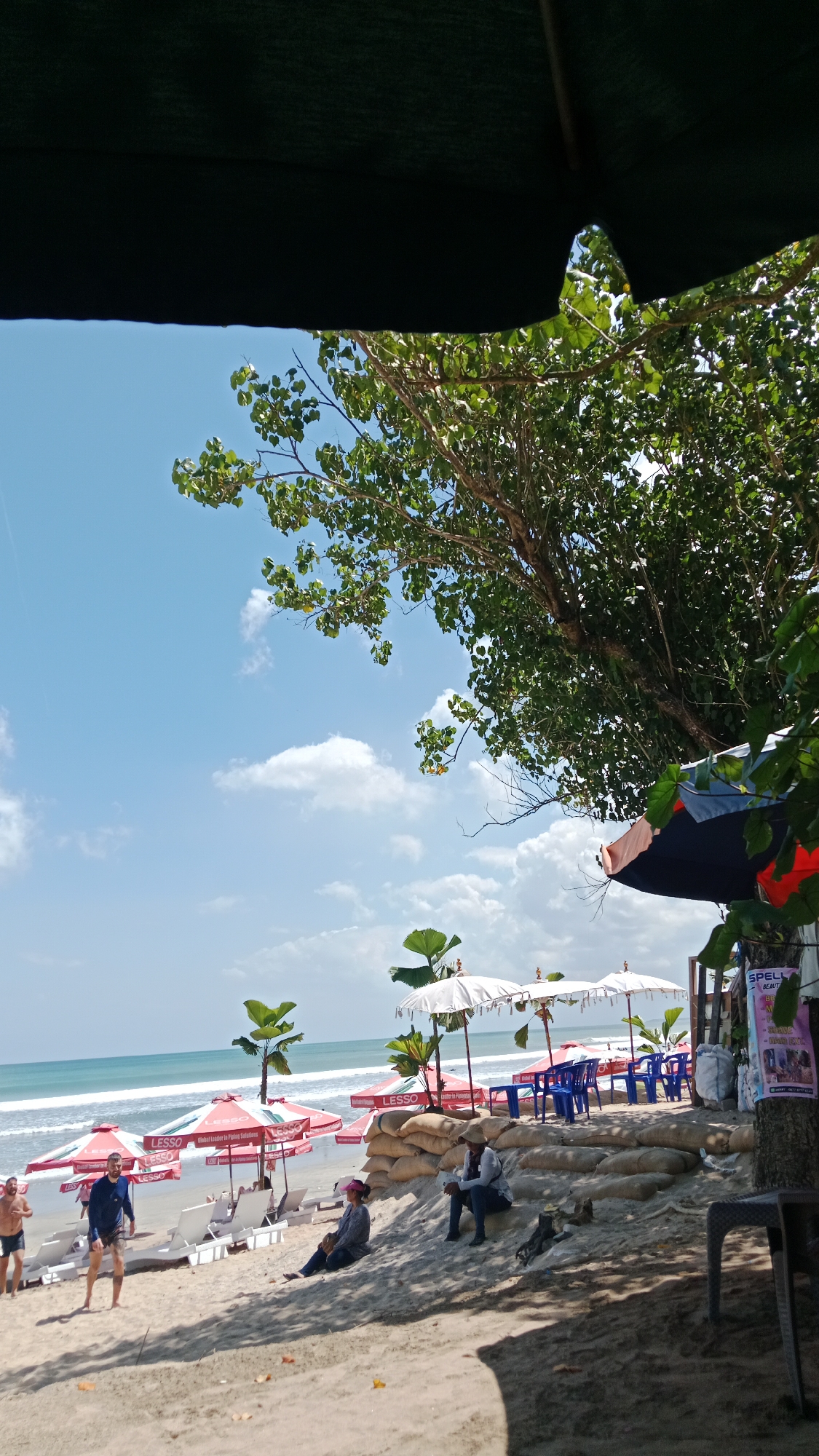 The image depicts a sunny day at a beach in Kuta, Indonesia, with the ocean visible in the background. The foreground is dominated by a sandy beach with several beach umbrellas, lounge chairs, and tables set up. A large tree with green leaves provides shade in the upper right portion of the frame.
Several people are visible in the scene. On the left, two men are standing on the beach. One is wearing a blue long-sleeved shirt and dark shorts, while the other is in black shorts. Further back, a person is lying on a lounge chair under a red umbrella.
In the mid-ground, a group of people are seated on sandbags. Two individuals are sitting on a sandbag wall, with one wearing a hat and a light-colored shirt, and the other wearing a dark shirt and jeans. Blue plastic chairs and tables are arranged behind them. There are several white parasols with decorative tops, and red umbrellas with the logo "LESSO" visible. Two young palm trees stand behind the red umbrellas.
On the far right, a tree trunk is visible with posters and signs attached to it. One sign appears to be for "SPELL BEAUTY." A dark plastic table and chair are in the lower right corner. The sky is blue with scattered white clouds. The overall impression is a recreational beach area in a tropical location.